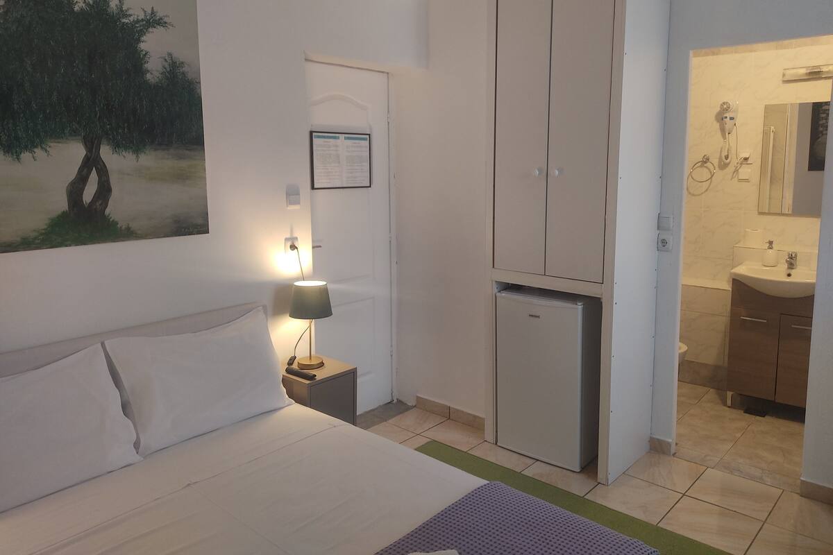 A hotel room with a bed, a nightstand with a lamp, a small refrigerator, a wardrobe, and a bathroom visible through an open door.