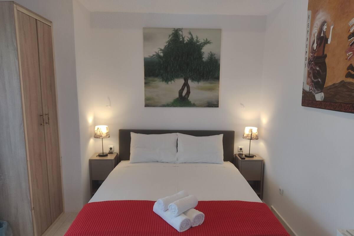 A neatly made bed with white linens and a red accent, two bedside lamps, a painting of a tree, and a wardrobe.