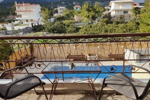A balcony with a glass table and chairs, overlooking a swimming pool and a residential area with houses and greenery. - Guest House Kithairon (Mandra-Eidyllia)