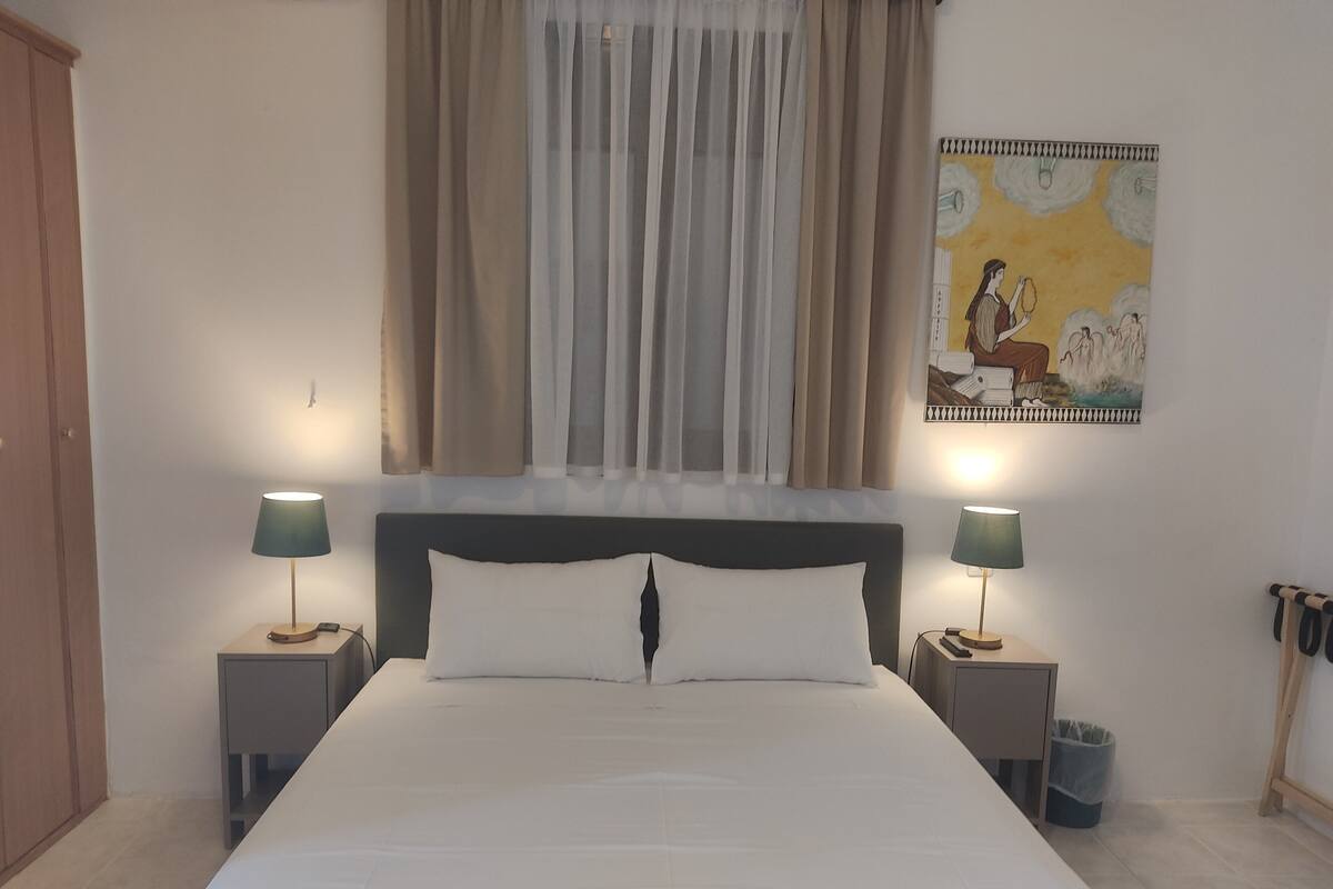 A neatly made bed with white linens, two bedside lamps, a headboard, a window with sheer curtains, and a wall-mounted artwork.