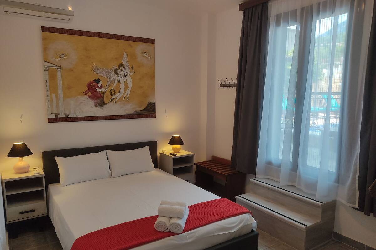 A hotel room with a large bed, two bedside tables with lamps, a window with curtains, and a painting on the wall.