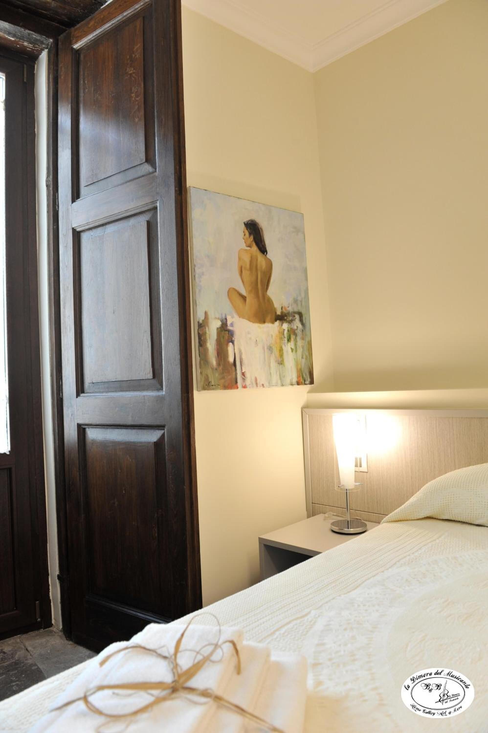 A hotel room with a large wooden door, a painting on the wall, a bedside table with a lamp, and a neatly made bed.