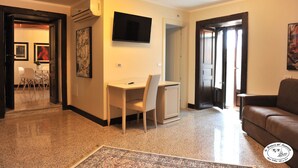 Een woonkamer met een bank, een aan de muur bevestigde televisie, een bureau met stoel, en een deur die naar een andere kamer leidt.