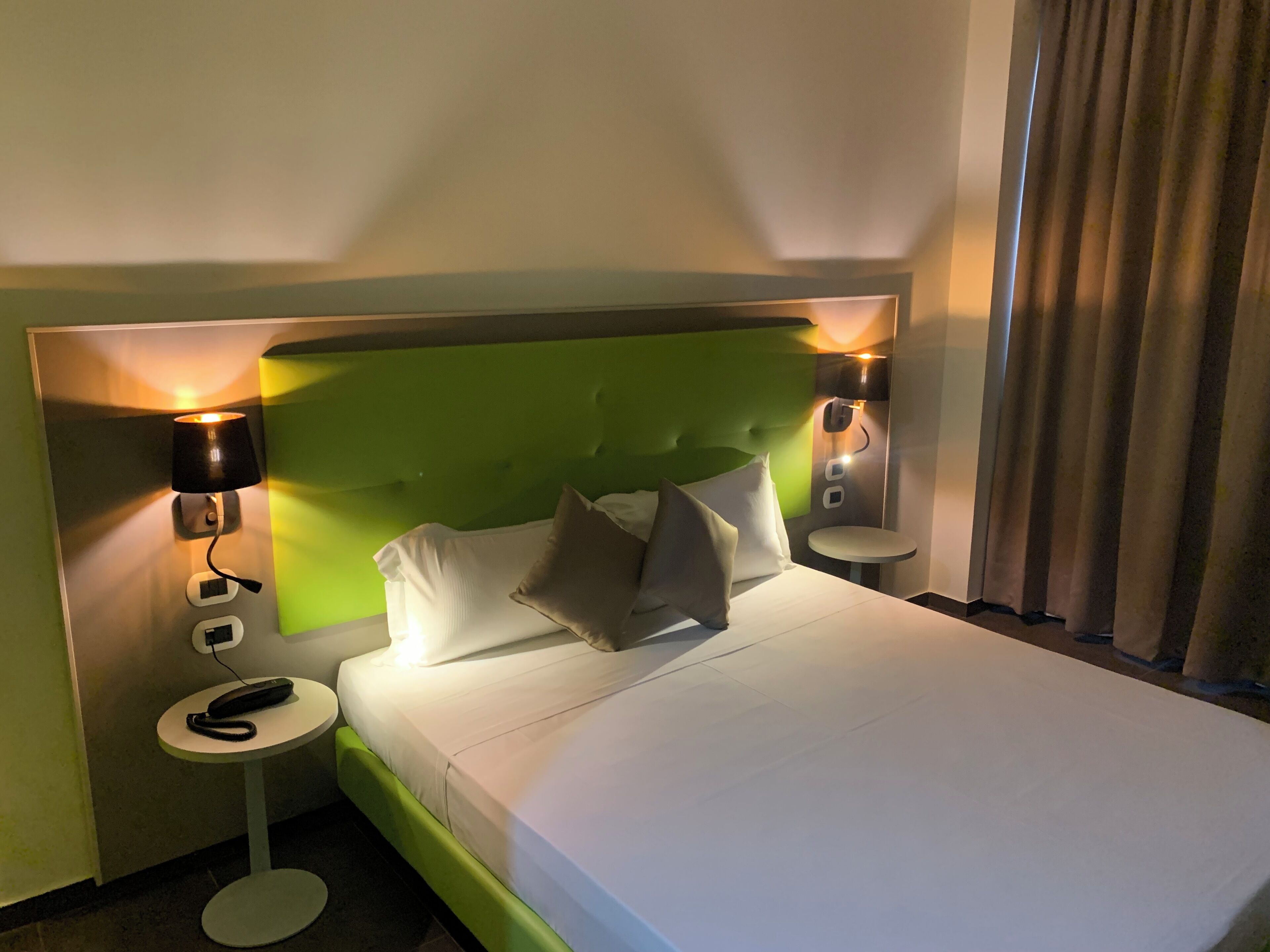 A hotel room with a green upholstered headboard, white bedding, two bedside lamps, and a small round table with a phone.