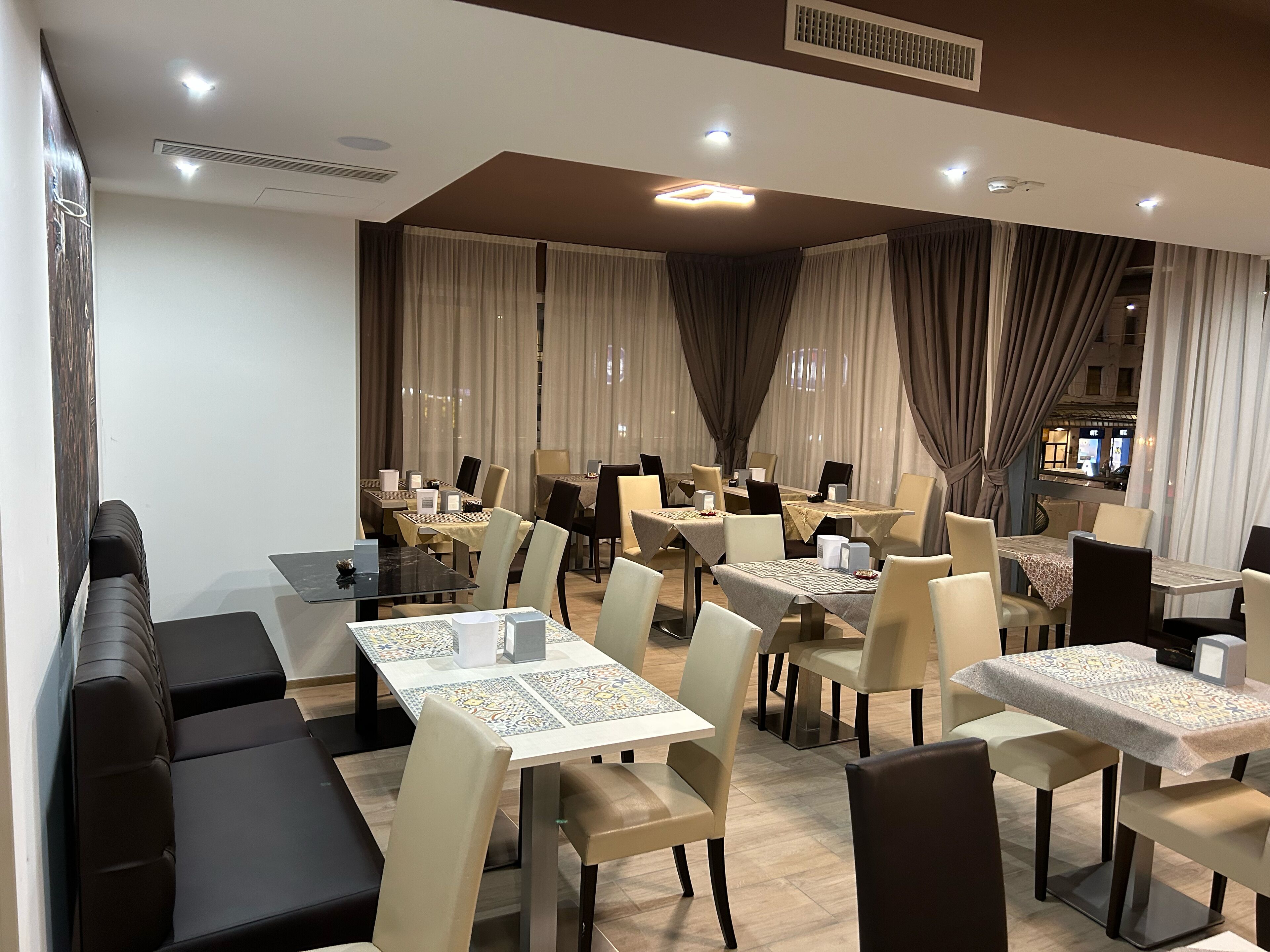 Un'elegante sala da pranzo con diversi tavoli e sedie, un divano e vista sulla strada attraverso ampie finestre.