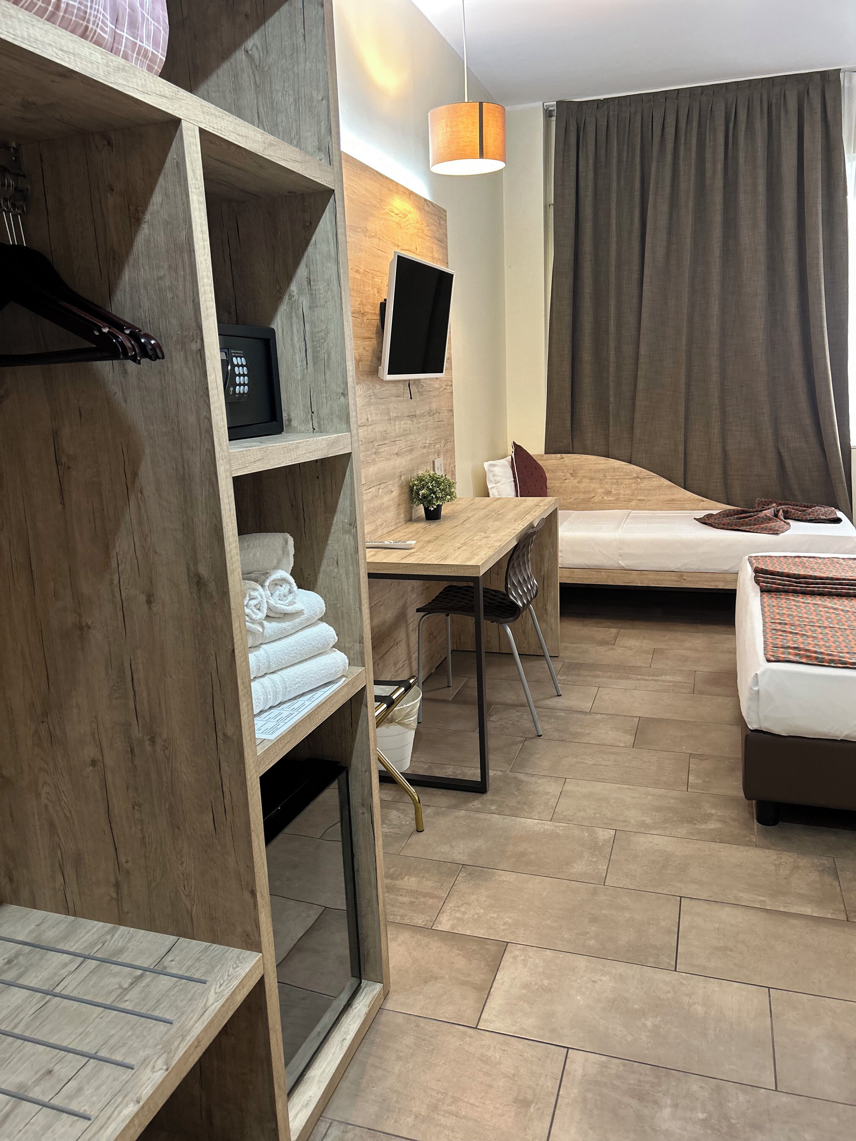 Une chambre d’hôtel avec une armoire en bois, un bureau avec une chaise, un lit, un téléviseur fixé au mur et une grande fenêtre avec des rideaux.