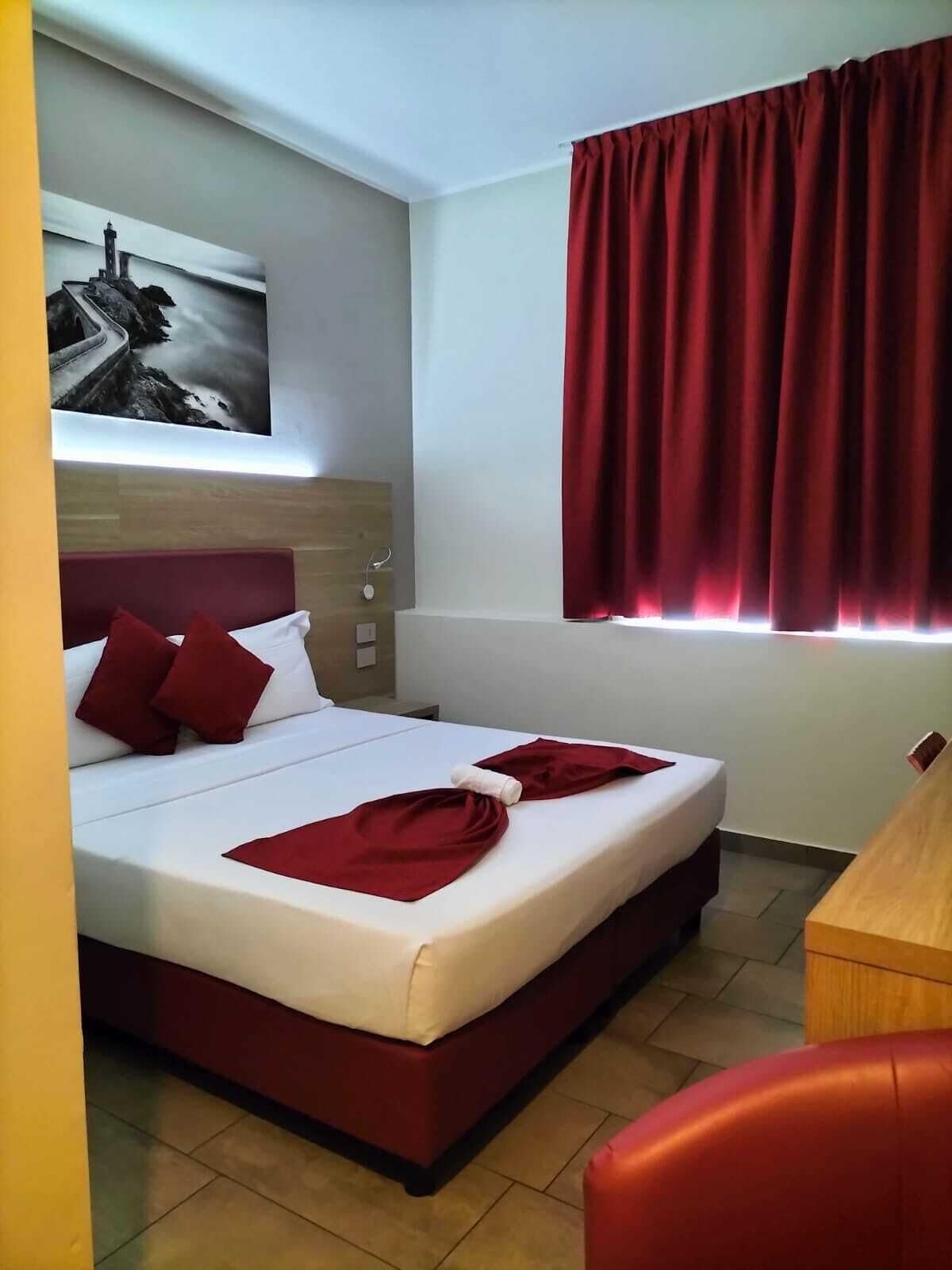 A hotel room with a bed, red curtains, a painting on the wall, and a wooden headboard.