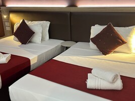 Habitación de hotel con dos camas, cada una con ropa blanca y un almohadón marrón. Hay una mesita redonda pequeña entre las camas, y un banco rojo al pie de cada cama.
