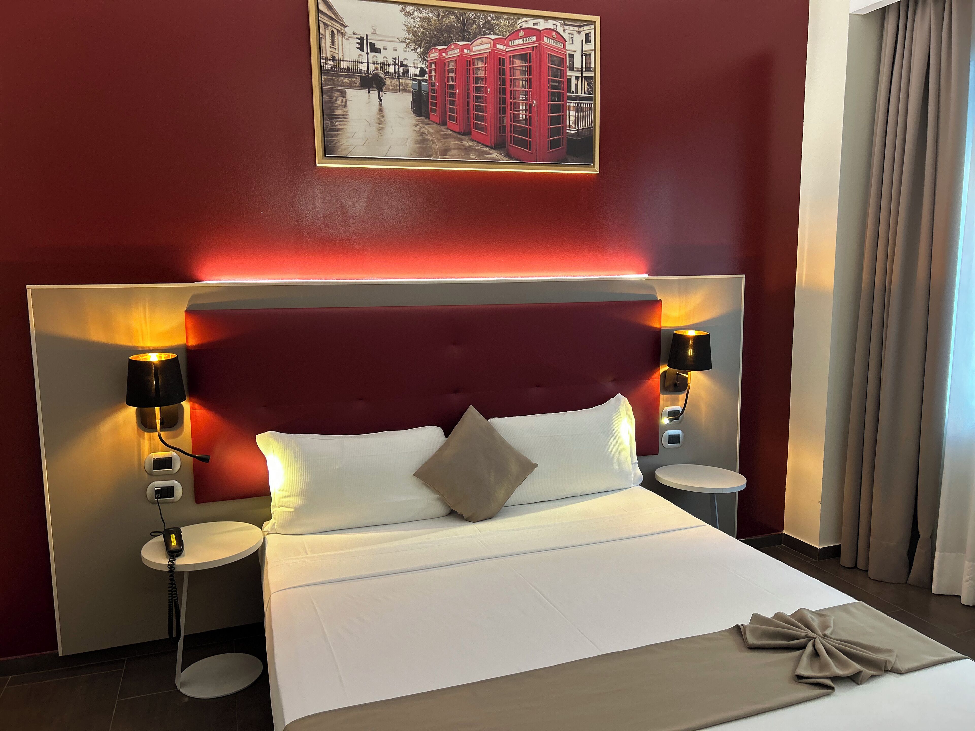 A hotel room with a large bed, two bedside tables, a headboard with integrated lighting, a wall-mounted picture, and a window with curtains.