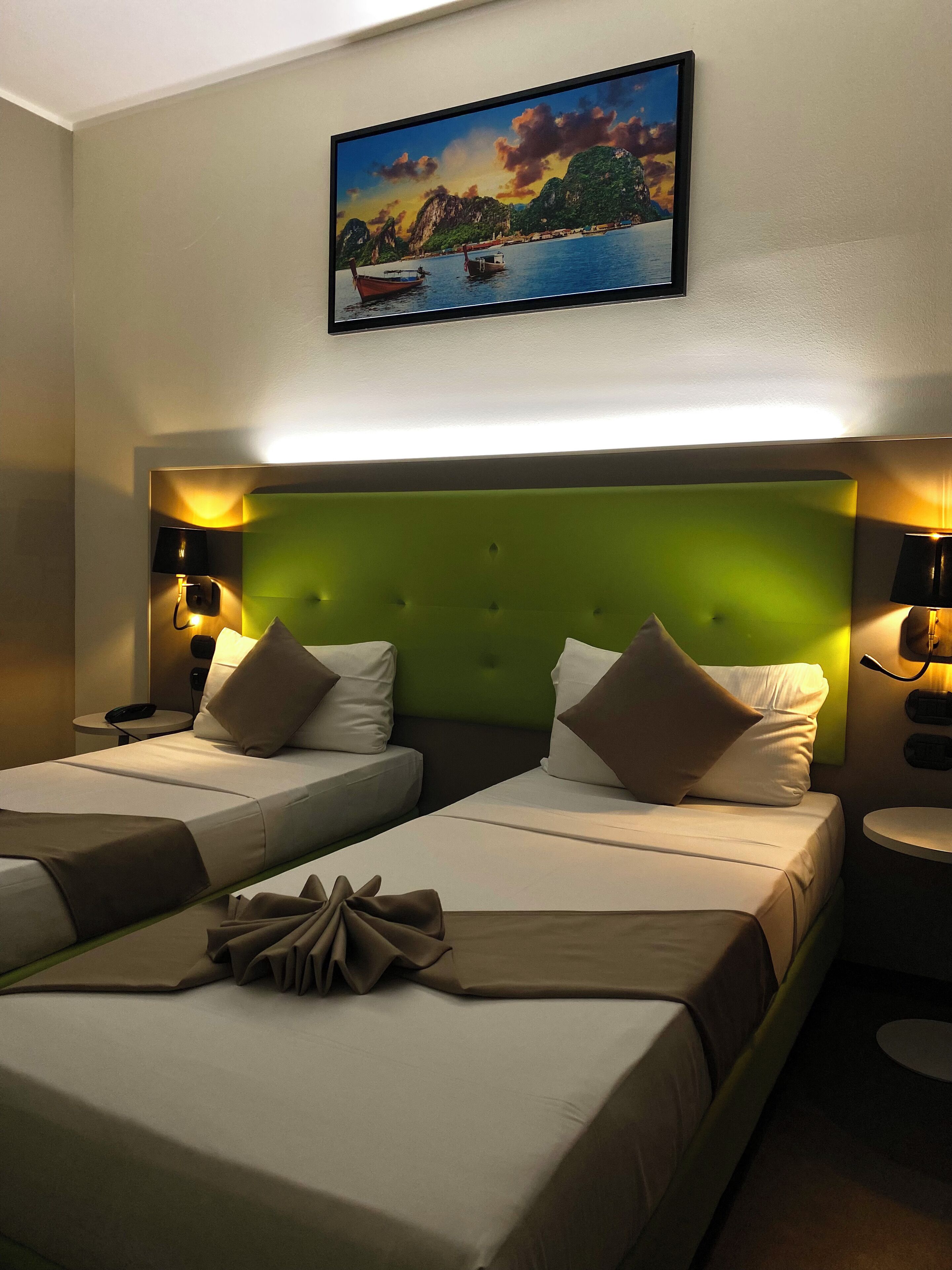 A hotel room with two beds, a green headboard, and a painting of a coastal scene above the beds.