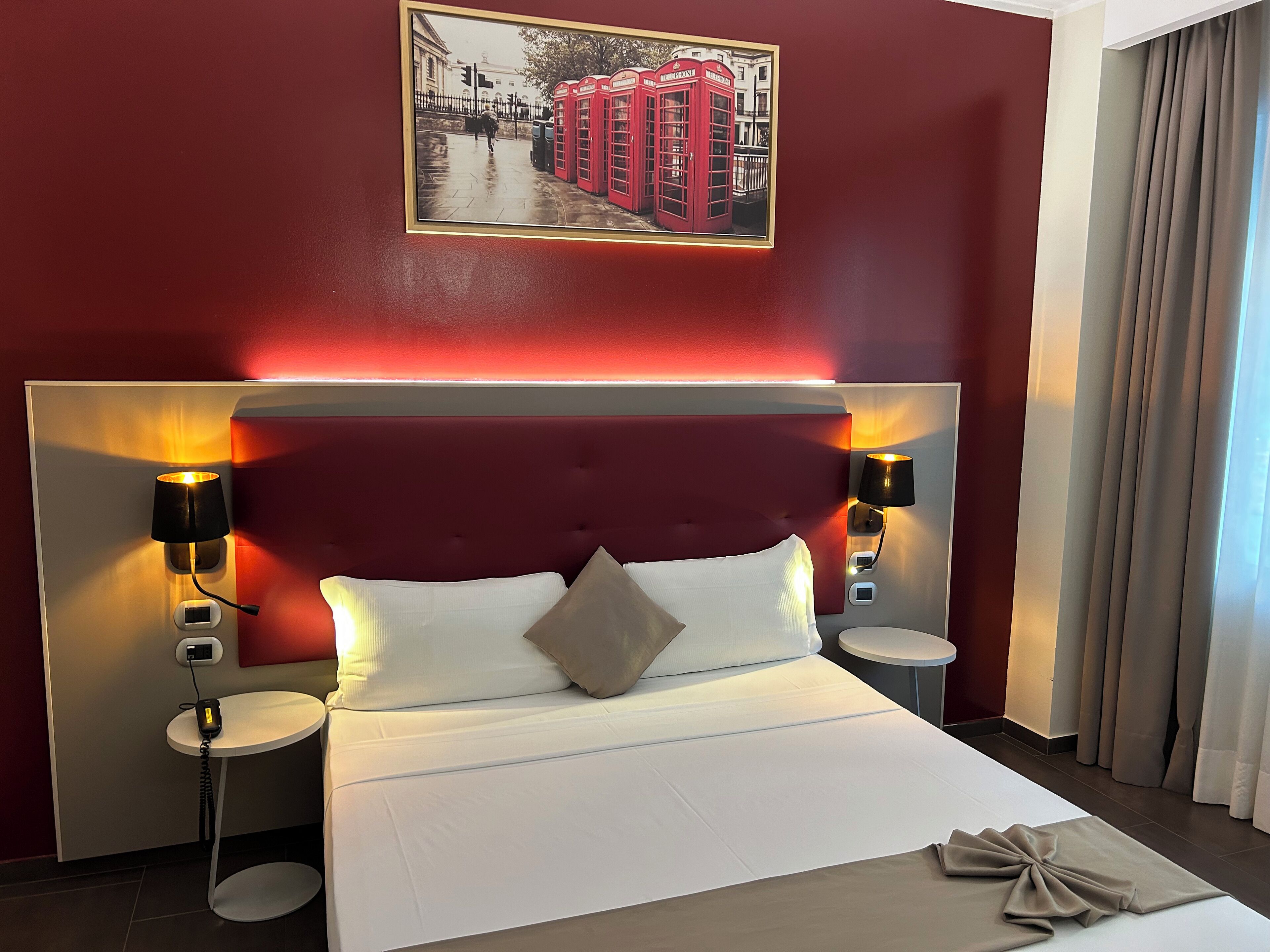 A hotel room with a large bed, two bedside tables, a headboard with integrated lighting, a wall-mounted lamp, and a framed picture above the bed.