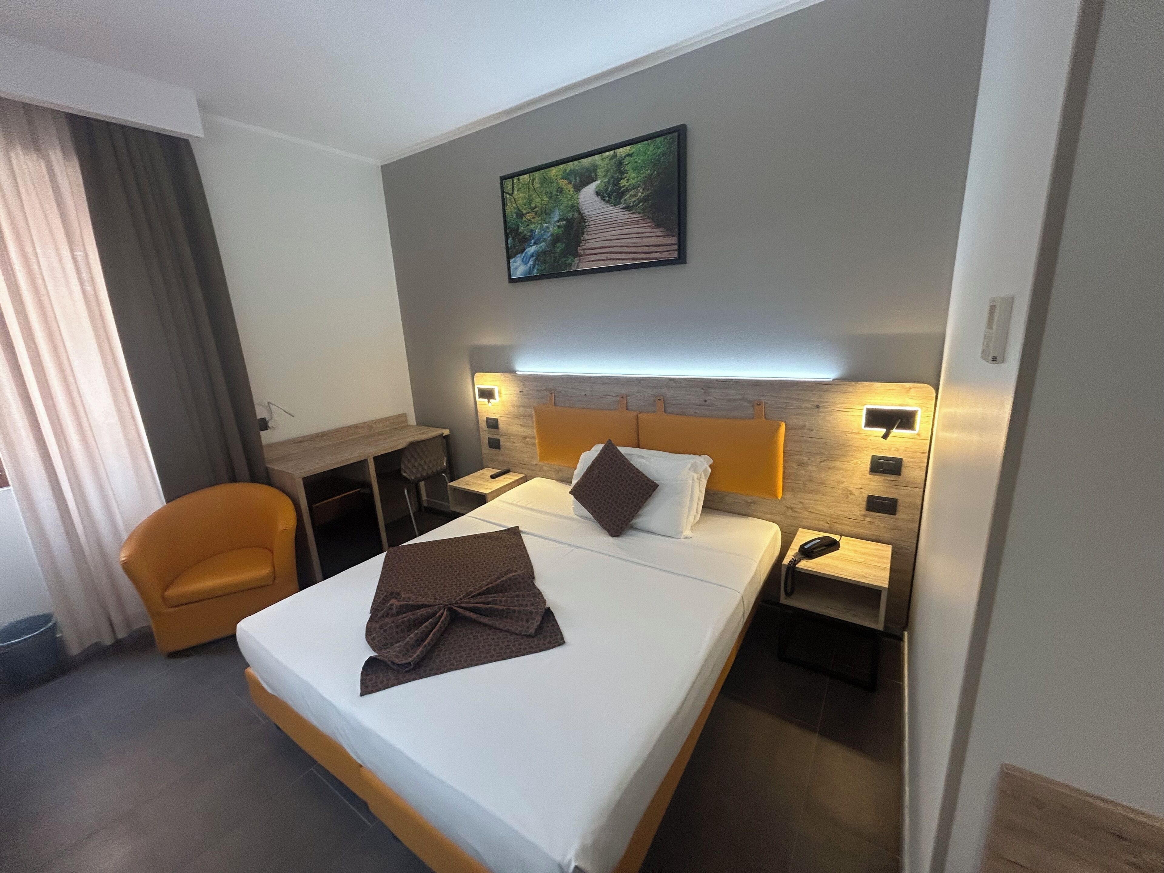 A hotel room with a bed, a desk, a chair, and a nightstand with a lamp.