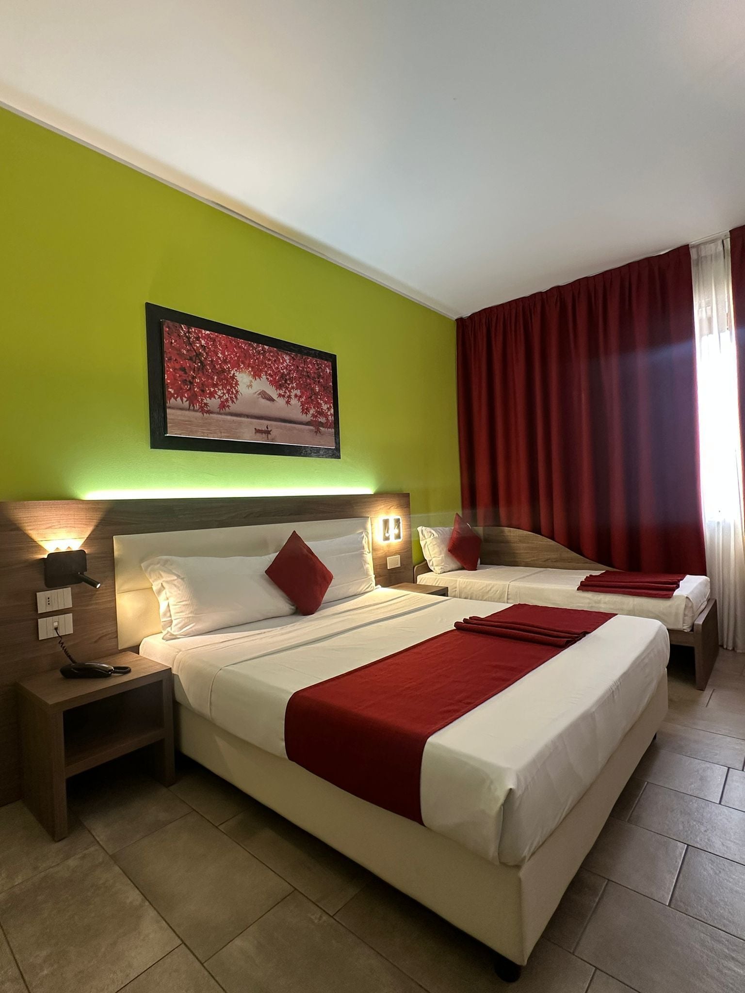Une chambre d’hôtel avec deux lits, une table de chevet avec un téléphone, une lampe fixée au mur et un tableau représentant un arbre rouge.