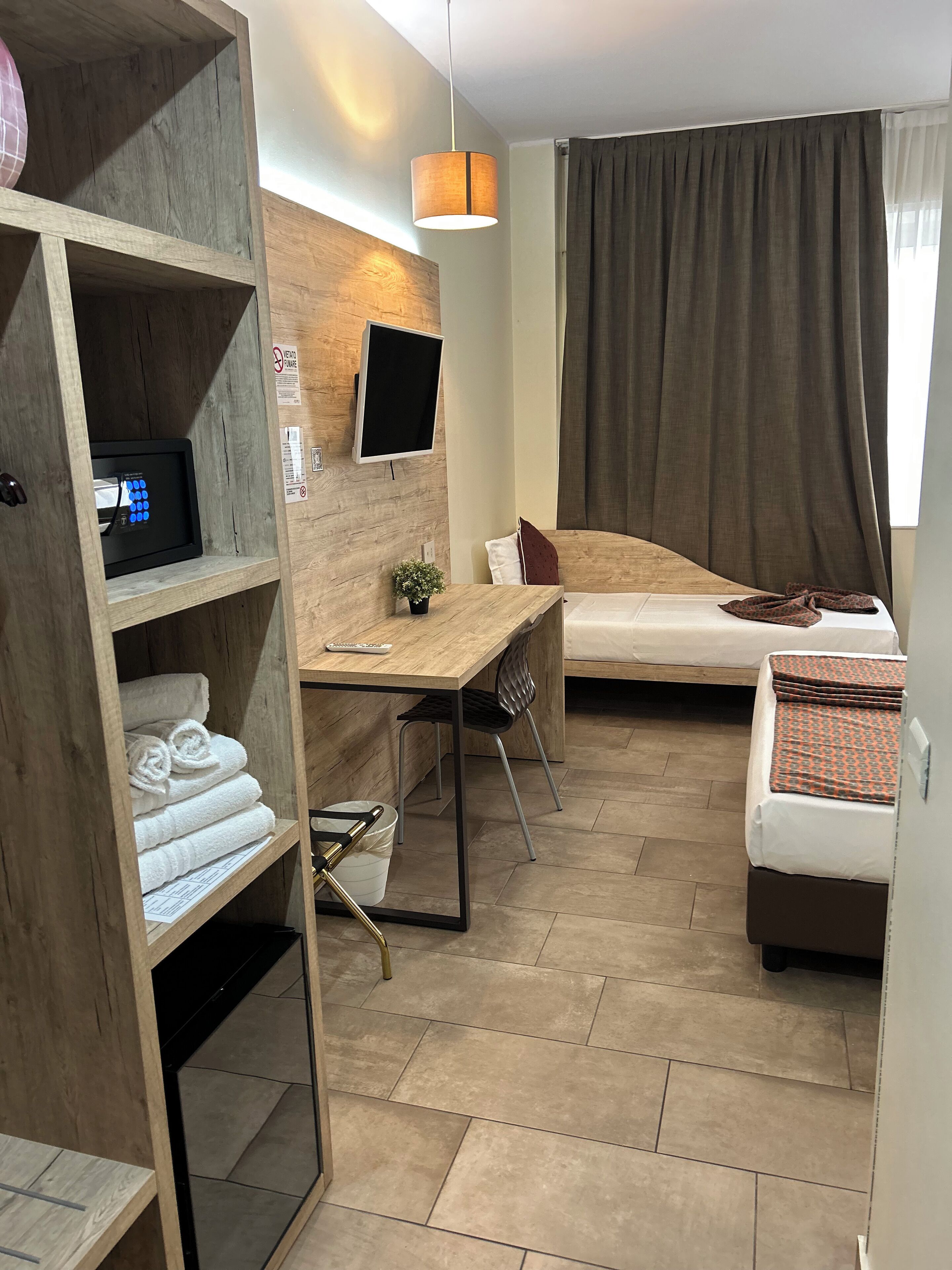 A compact hotel room with a wooden desk, a bed, a small seating area, a TV mounted on the wall, and a large window with curtains.
