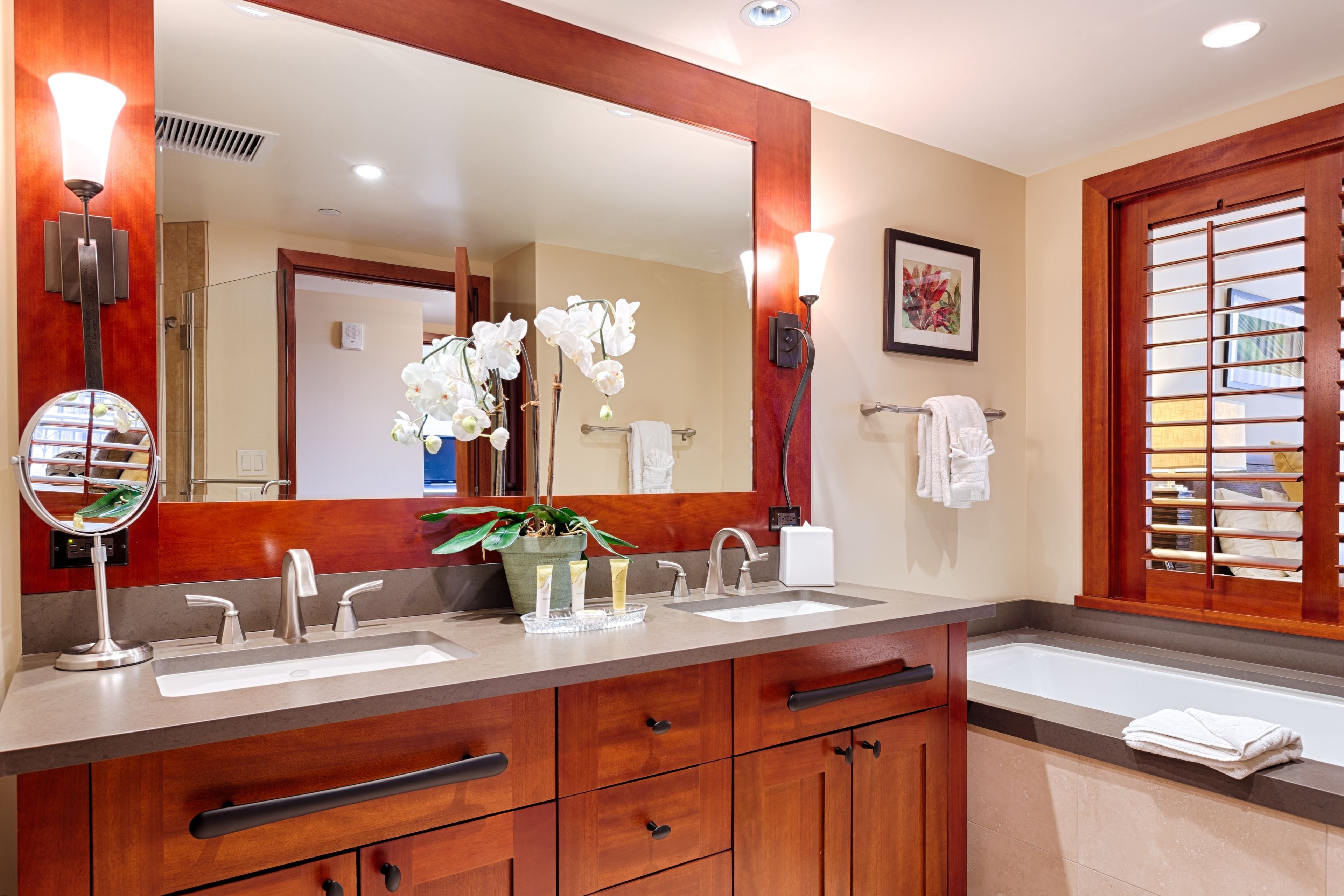 Un baño con un espejo grande, dos lavamanos, un mueble de tocador con cajones, una orquídea en maceta y un perchero para toallas.