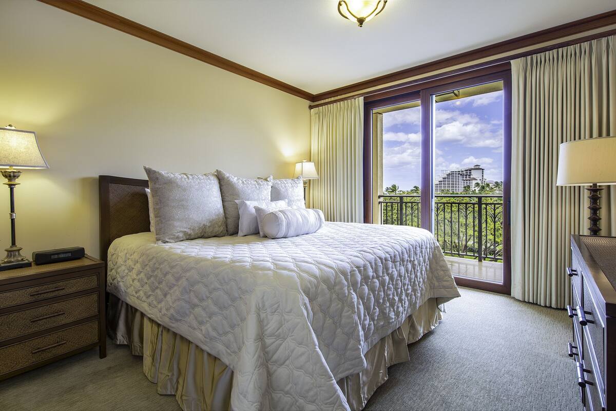Habitación de hotel con una cama grande, una mesita de noche, un aparador y un balcón con vistas a un paisaje urbano.