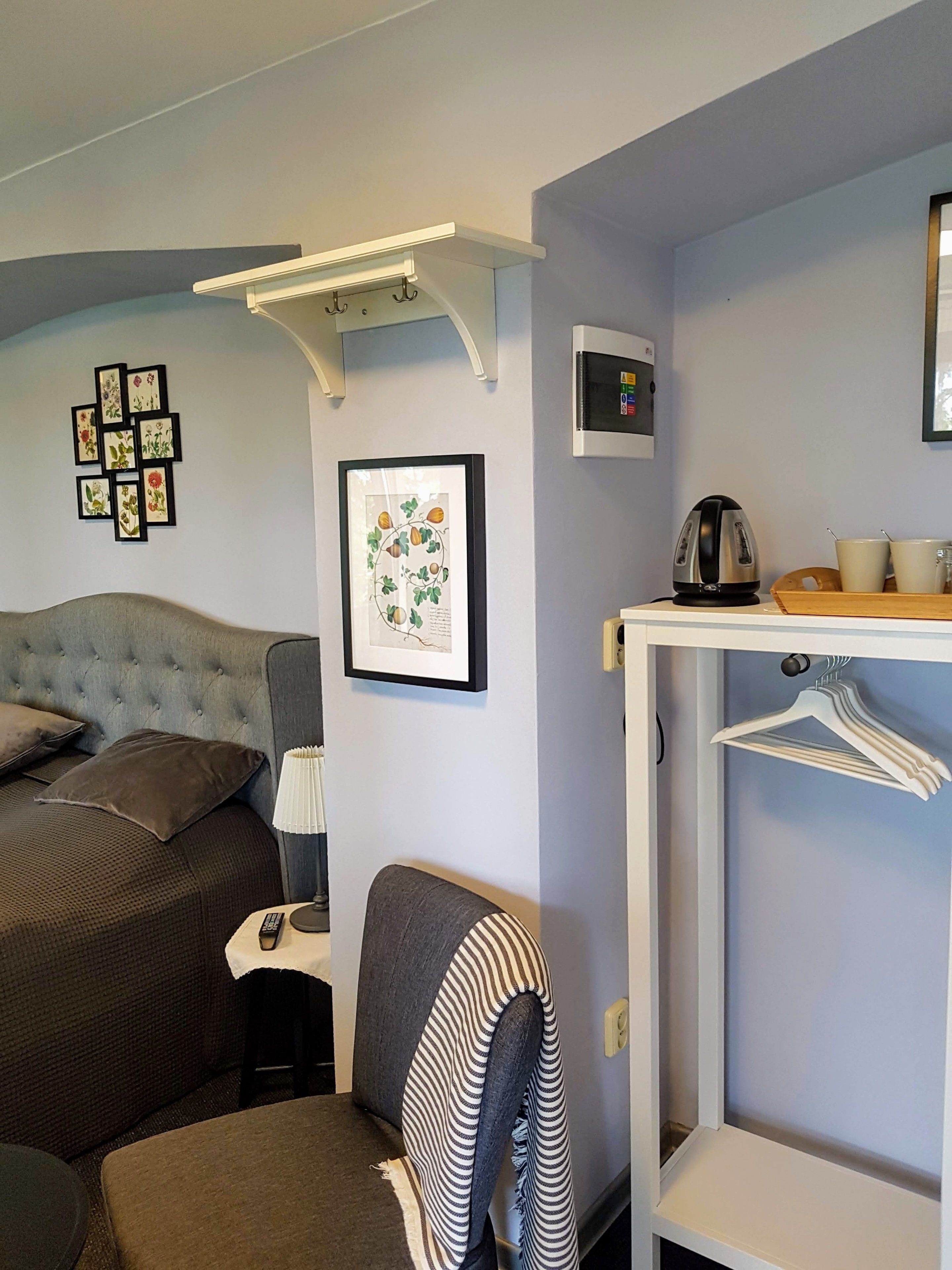 A neatly arranged hotel room with a bed, a chair, a table with a kettle, and a wall-mounted heater.