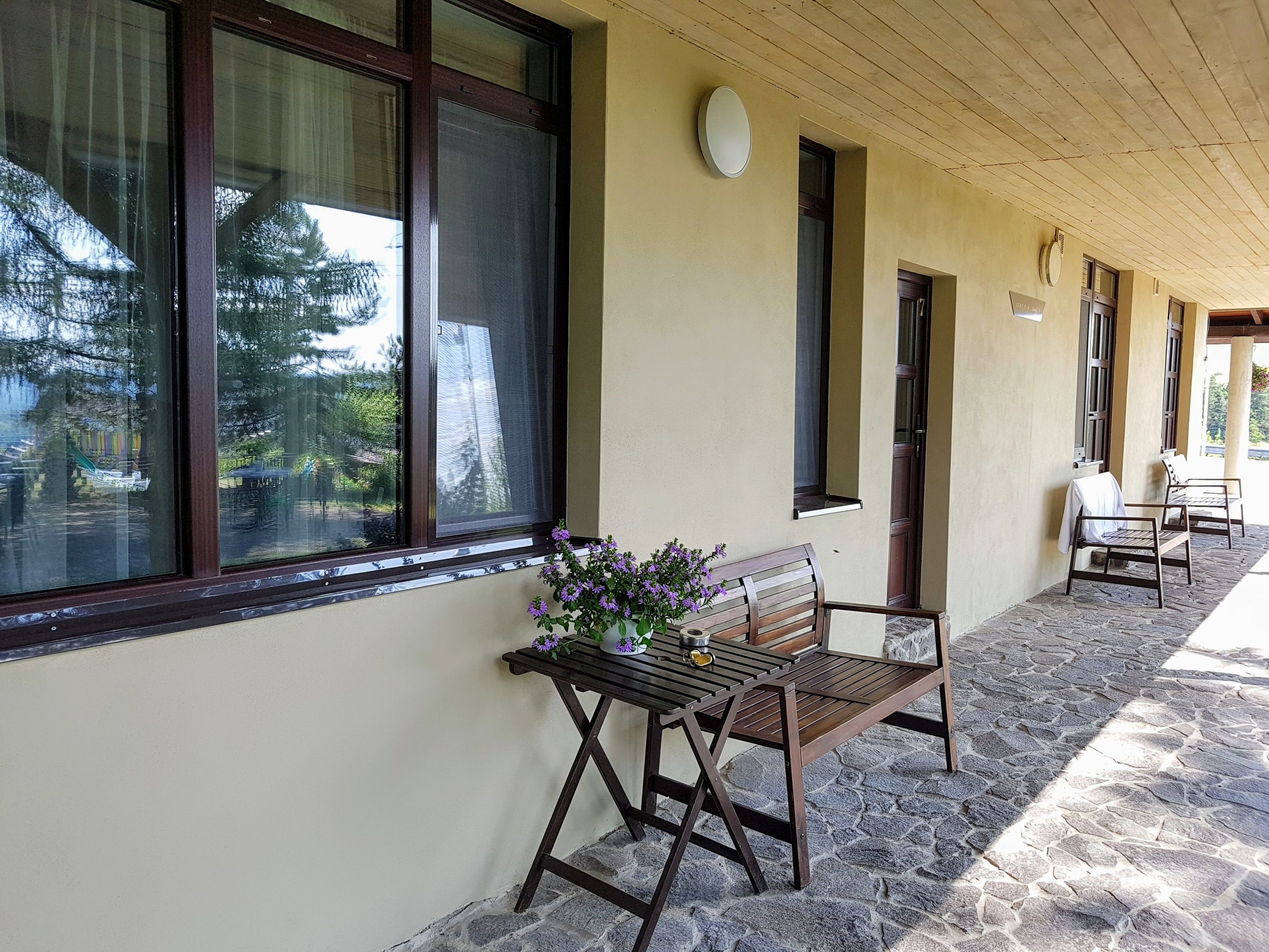 A covered outdoor area with a bench and a table, stone flooring, and windows with blinds.