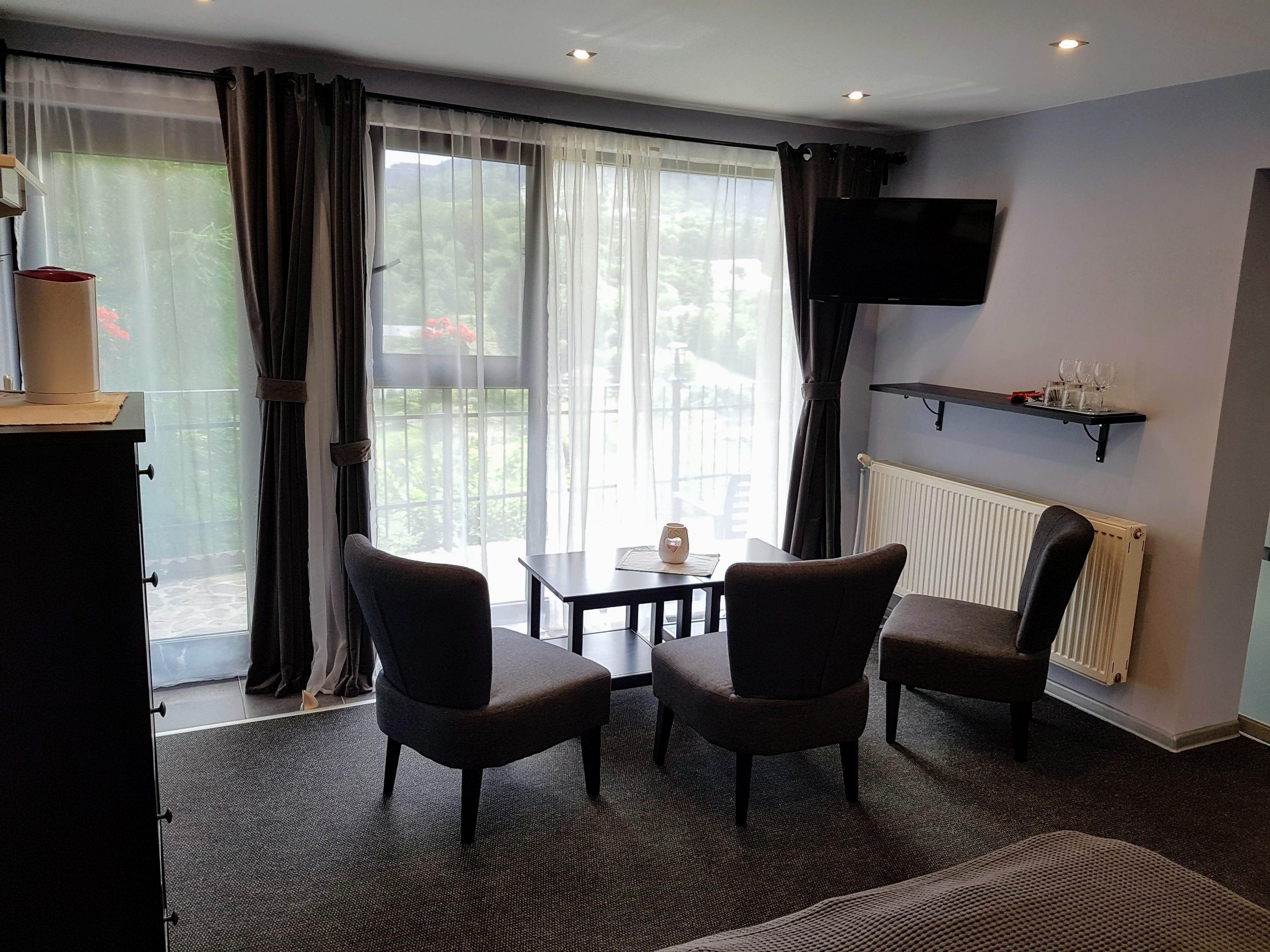 A room with a large window, a flat-screen TV mounted on the wall, a table with two chairs, and a radiator.