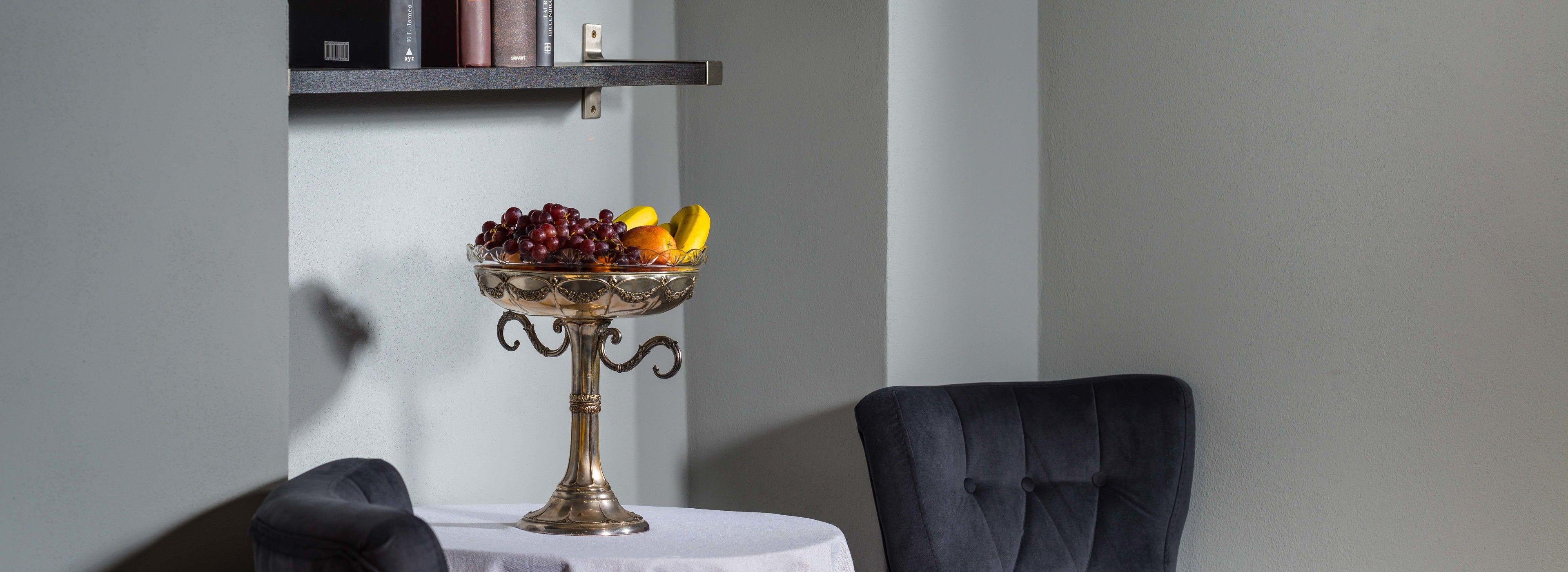 A table with a fruit bowl, a chair, and a bookshelf against a wall.