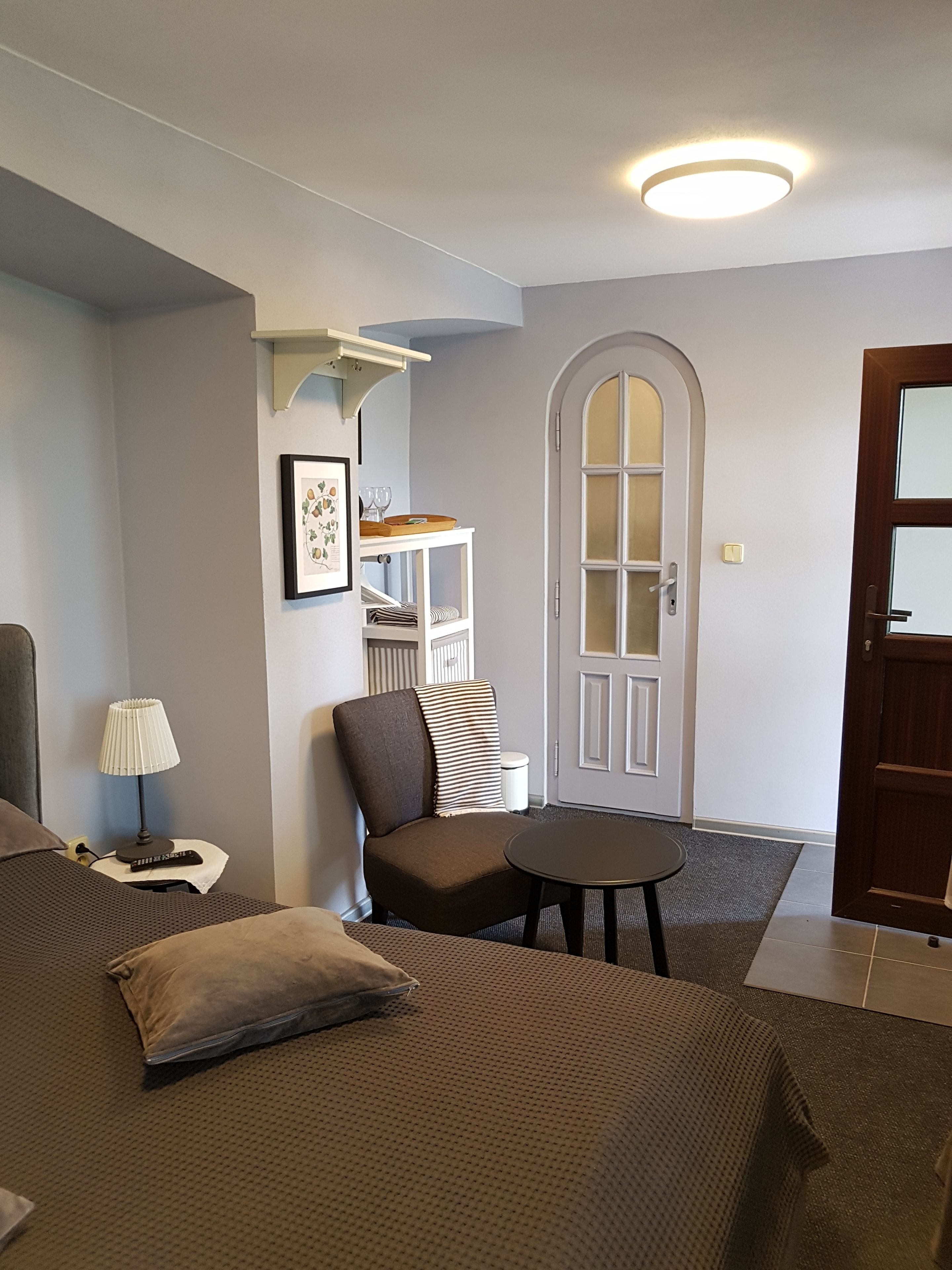 Une chambre d’hôtel avec un lit, une chaise, une petite table, une lampe, un tableau au mur et une porte.