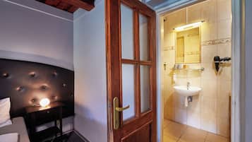 A hotel room with a wooden headboard, a bedside table with a lamp, and a door leading to a bathroom with a sink and mirror.
