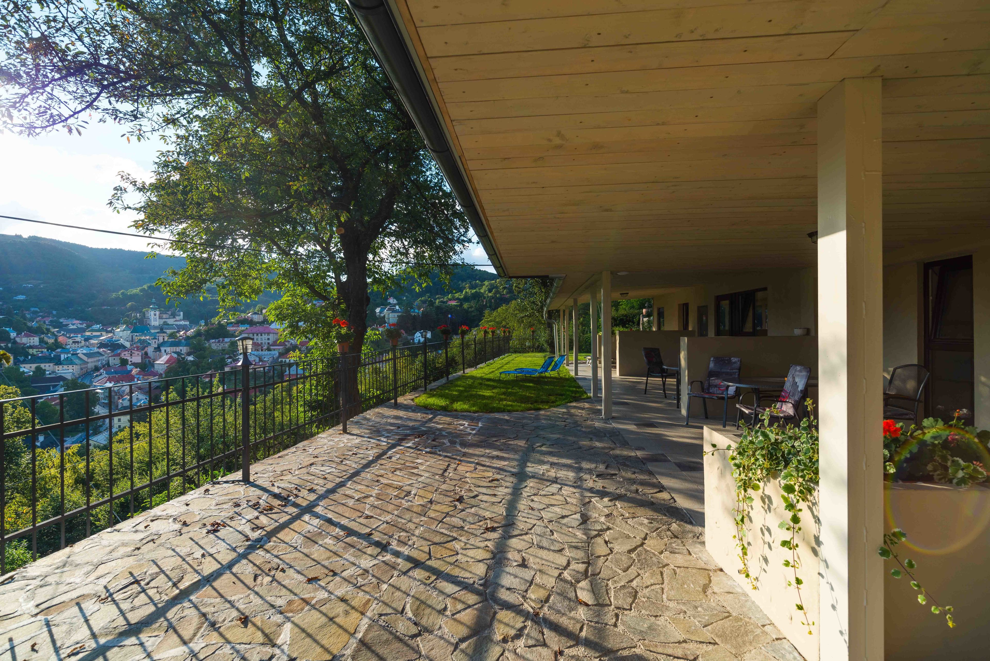A stone-paved balcony with a view of a town, a grassy area, and a seating area with chairs and a table.