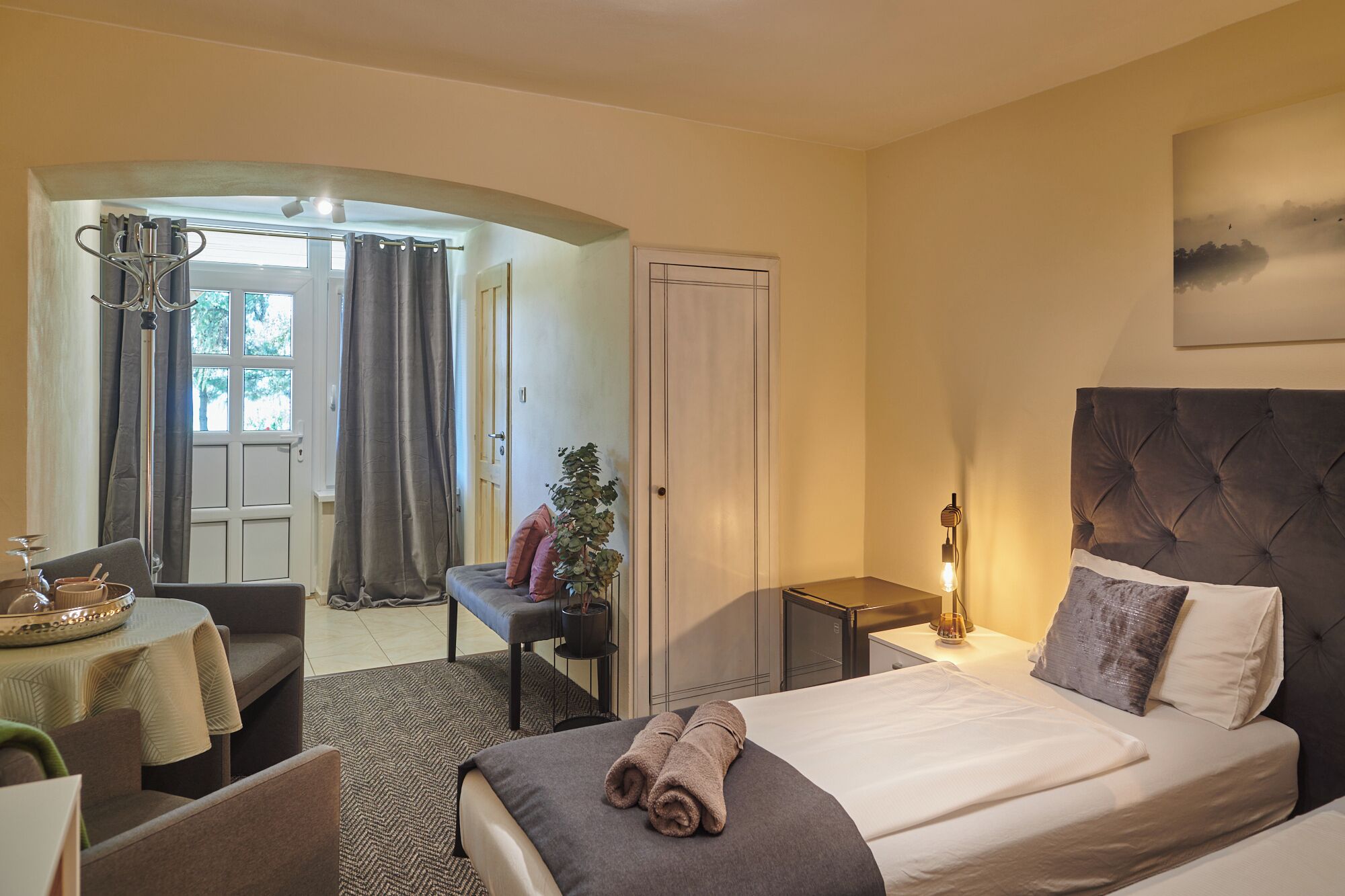 A hotel room with a bed, bedside table, lamp, and a view of the outside.