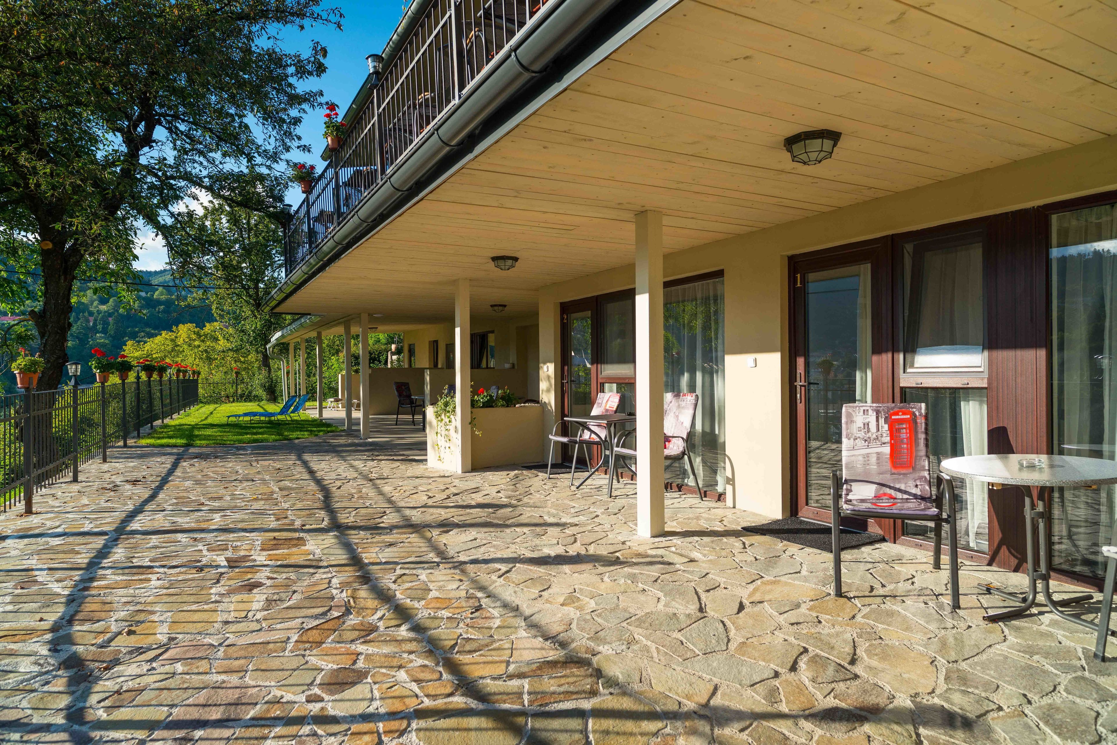 A stone-paved outdoor area with a covered seating area, a balcony with plants, and a view of the landscape.