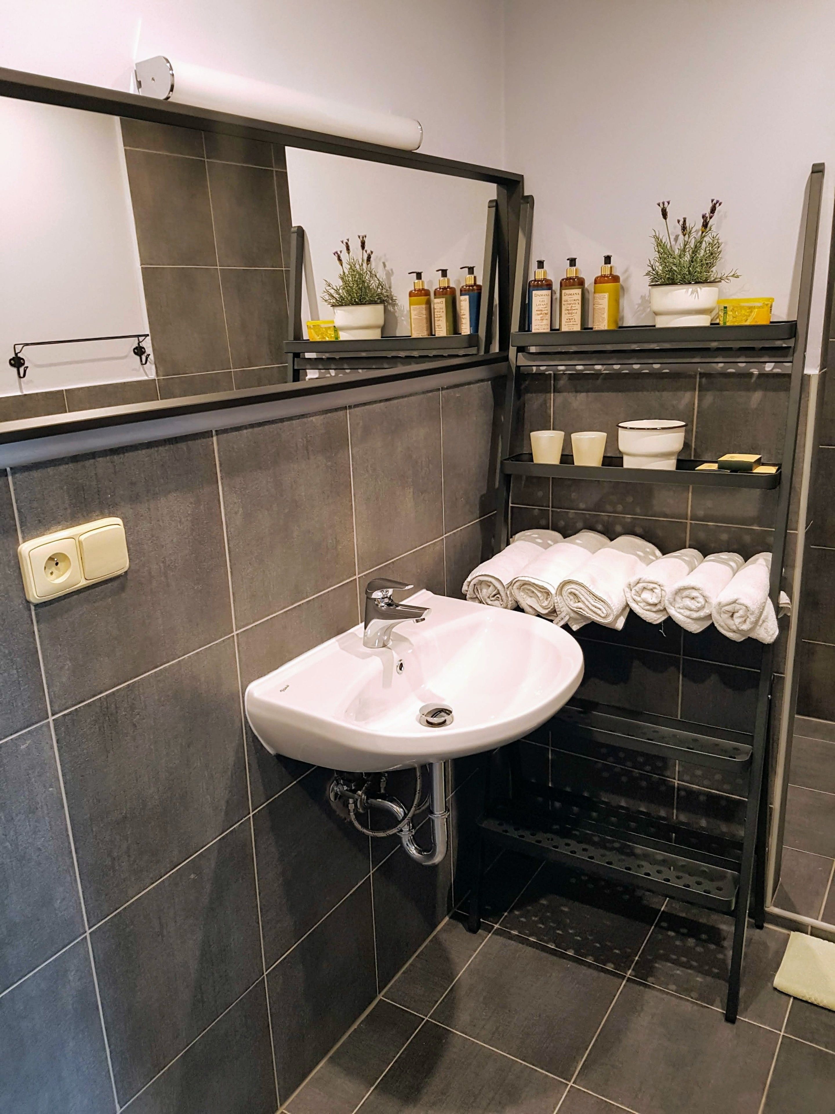 Et moderne badeværelse med en hvid vask, mørke flisevægge, et spejl og hylder med toiletartikler og håndklæder.