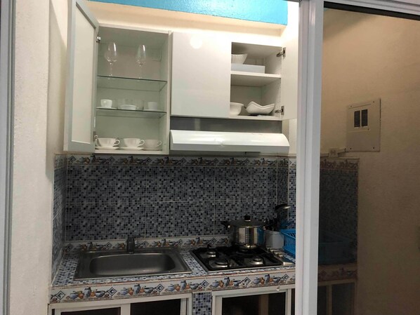A compact kitchen with a tiled backsplash, a sink, a stove, and open cabinets with dishes and glasses. - Aparta Hotel Azzurra (Boca Chica)