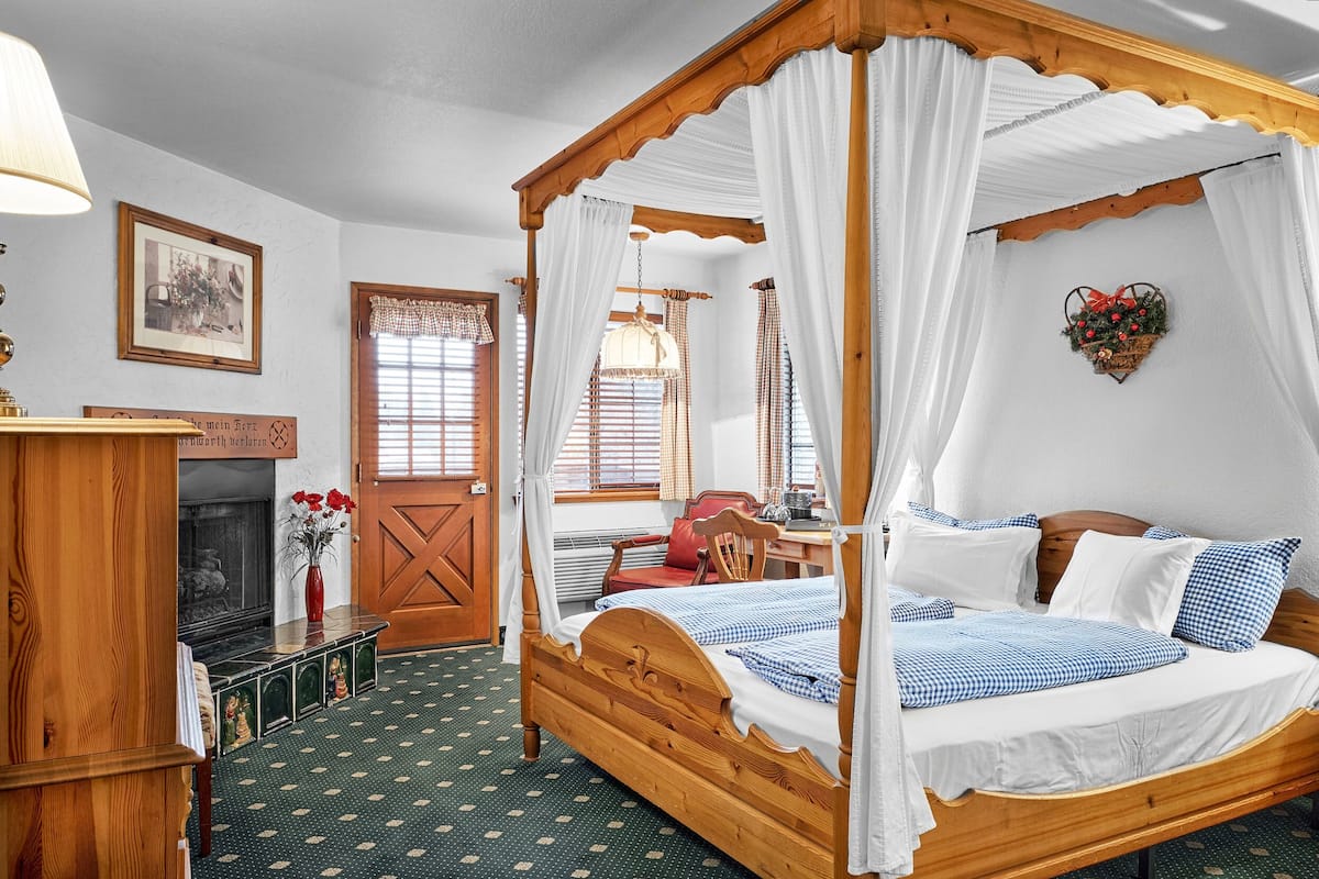 Een hemelbed met vier poten, wit beddengoed, een houten hoofdbord en een groen geweven vloerkleed.