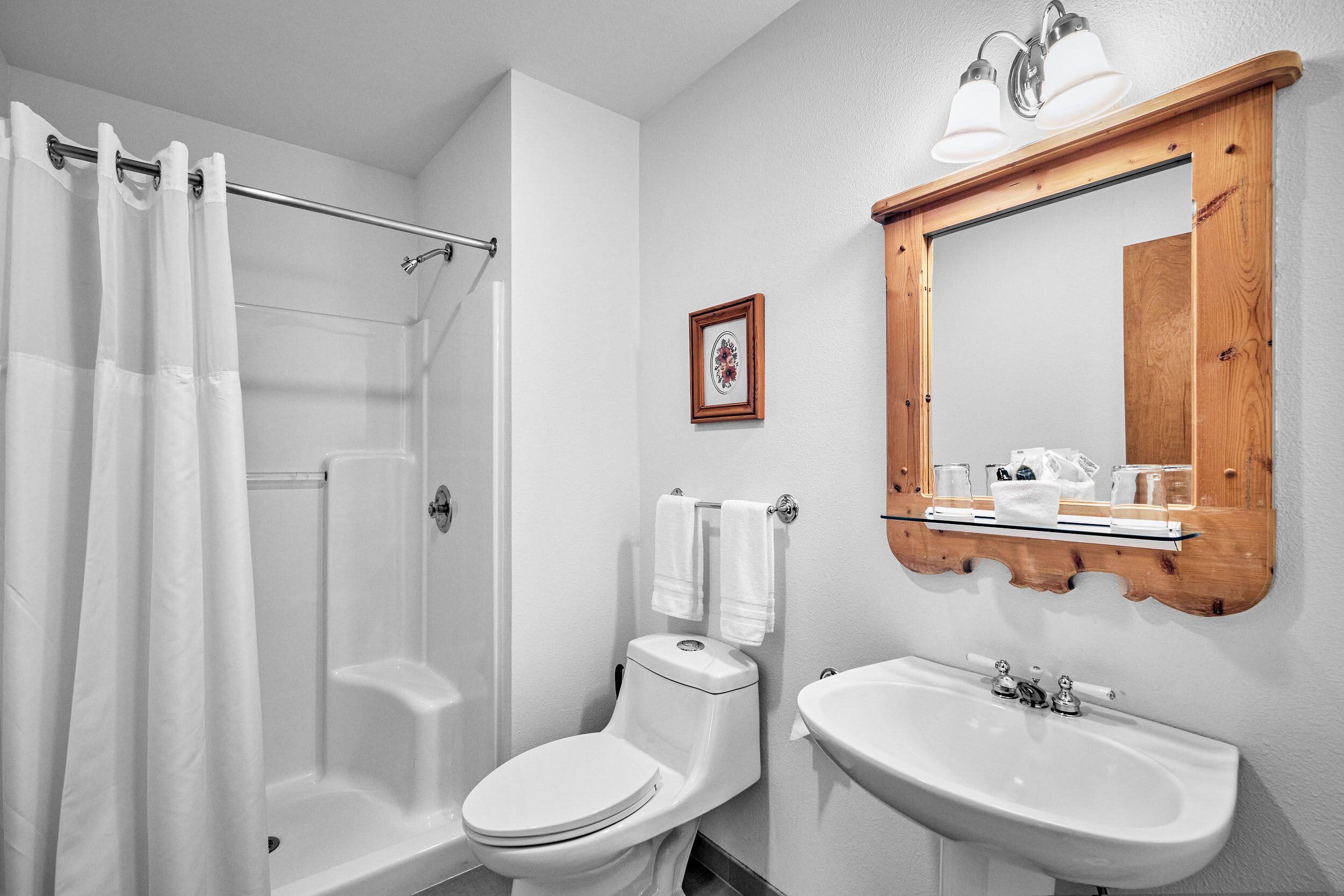 Ein Badezimmer mit Dusche, Toilette, Waschbecken und einem spiegelnden Wandbild mit Holzrahmen.