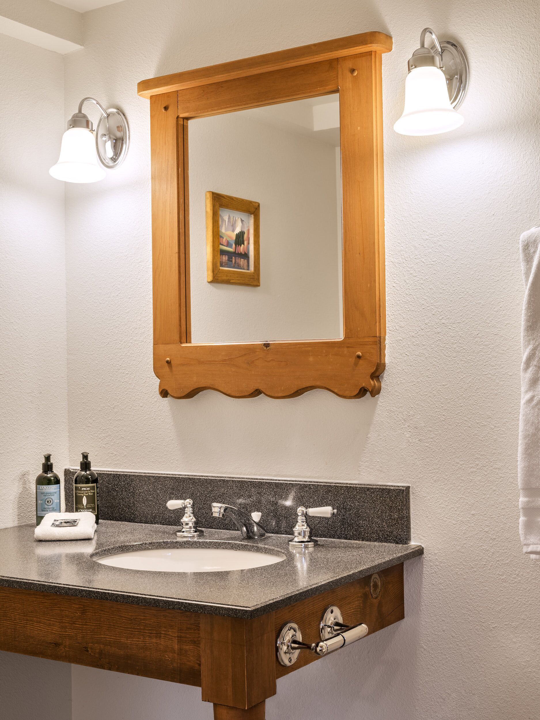 Ein Badezimmer mit einem spiegelnden Holzbettrahmen, zwei Wandleuchten, einer Granit-Arbeitsplatte, einem Waschbecken und einem Handtuchhalter.