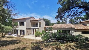 Um edifício de dois andares com varanda, alpendre e área de jardim com plantas e uma mesa pequena.