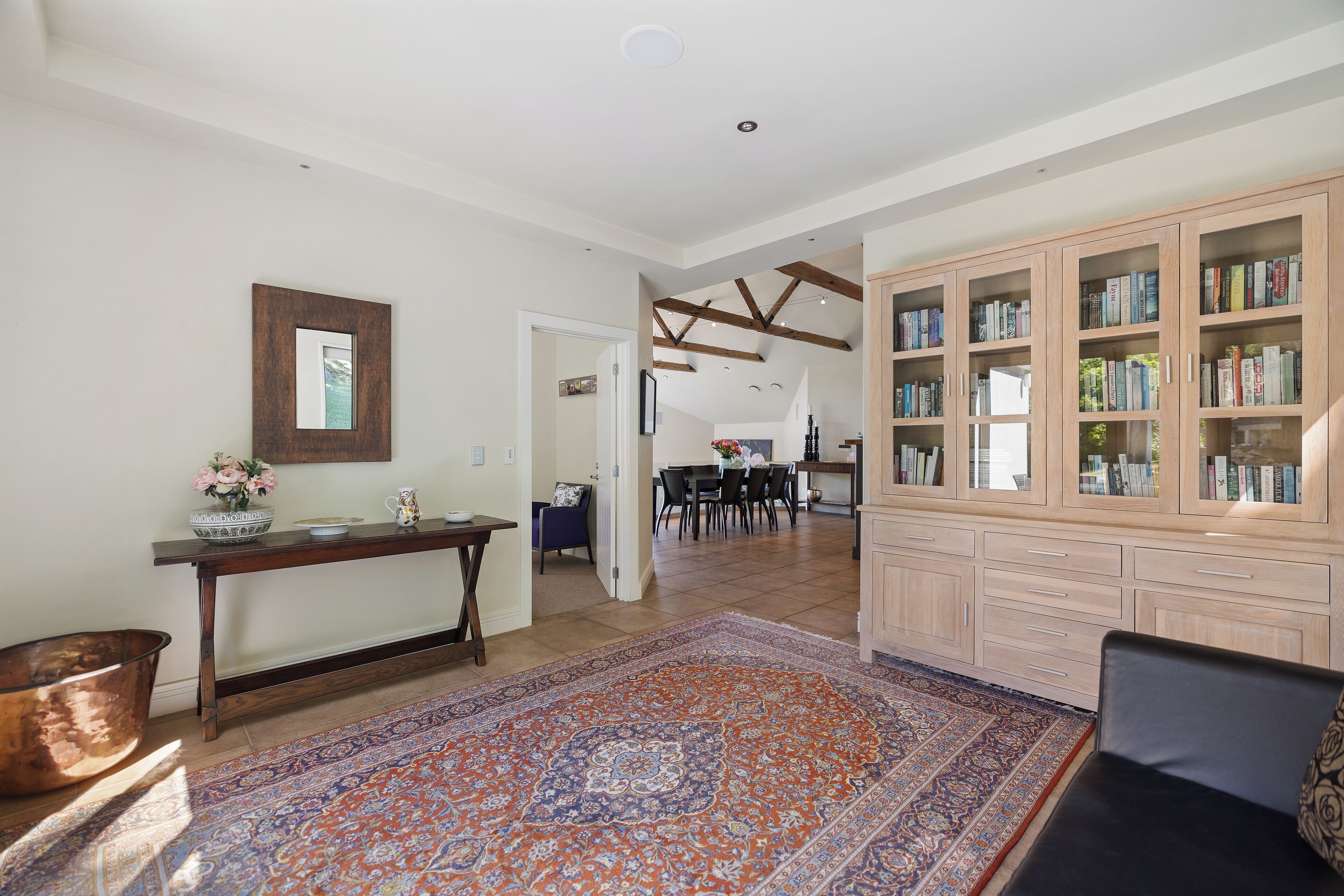 A spacious living area with a large wooden bookshelf filled with books, a dining area with a table and chairs, and a patterned rug on the floor.