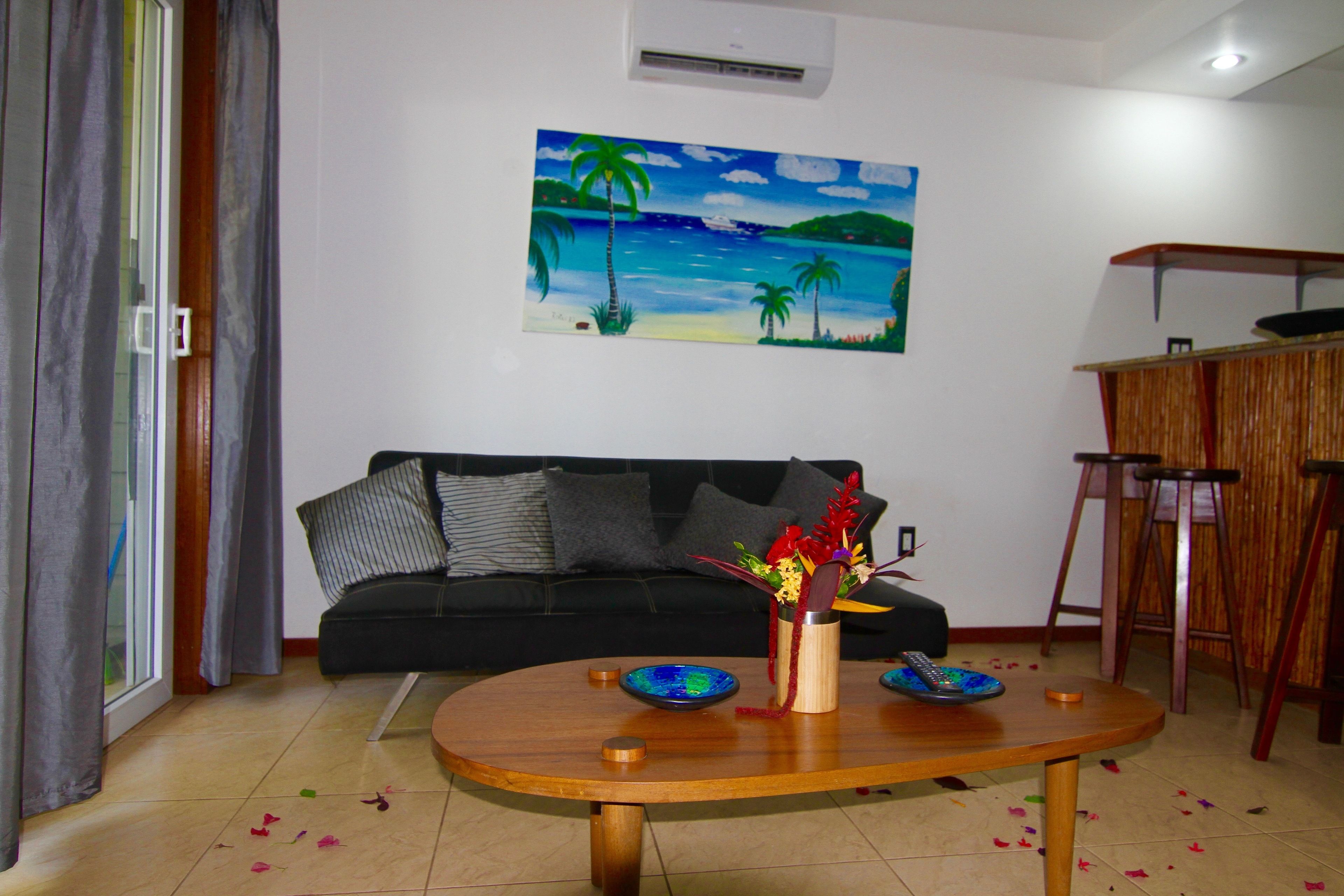 Ein Wohnzimmer mit einer schwarzen Couch, einem Holztisch mit Dekoration, einem Gemälde einer Strandansicht und einer kleinen Barzeile.