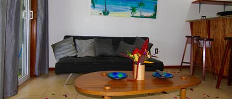 A living room with a black sofa, a wooden coffee table with decorative items, a painting of a beach scene, and a small bar area.