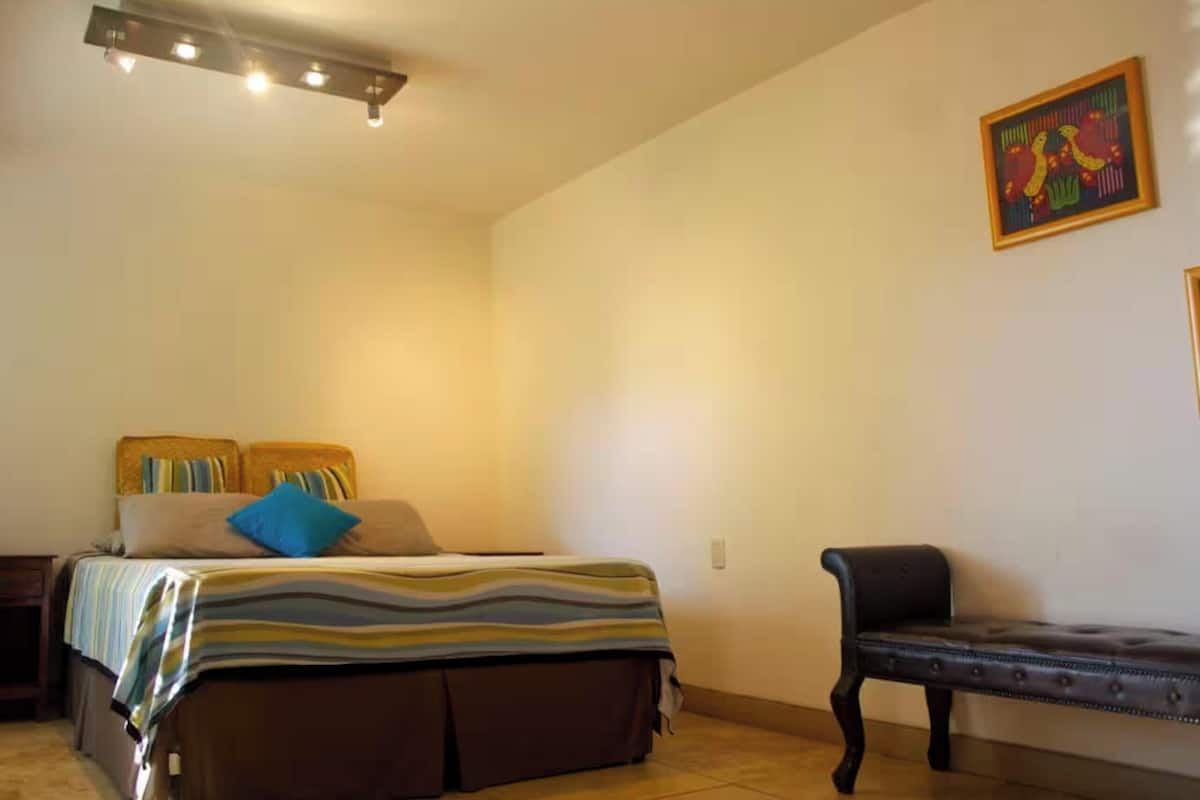 A bedroom with a bed, a bench, a wall-mounted light fixture, and a painting.