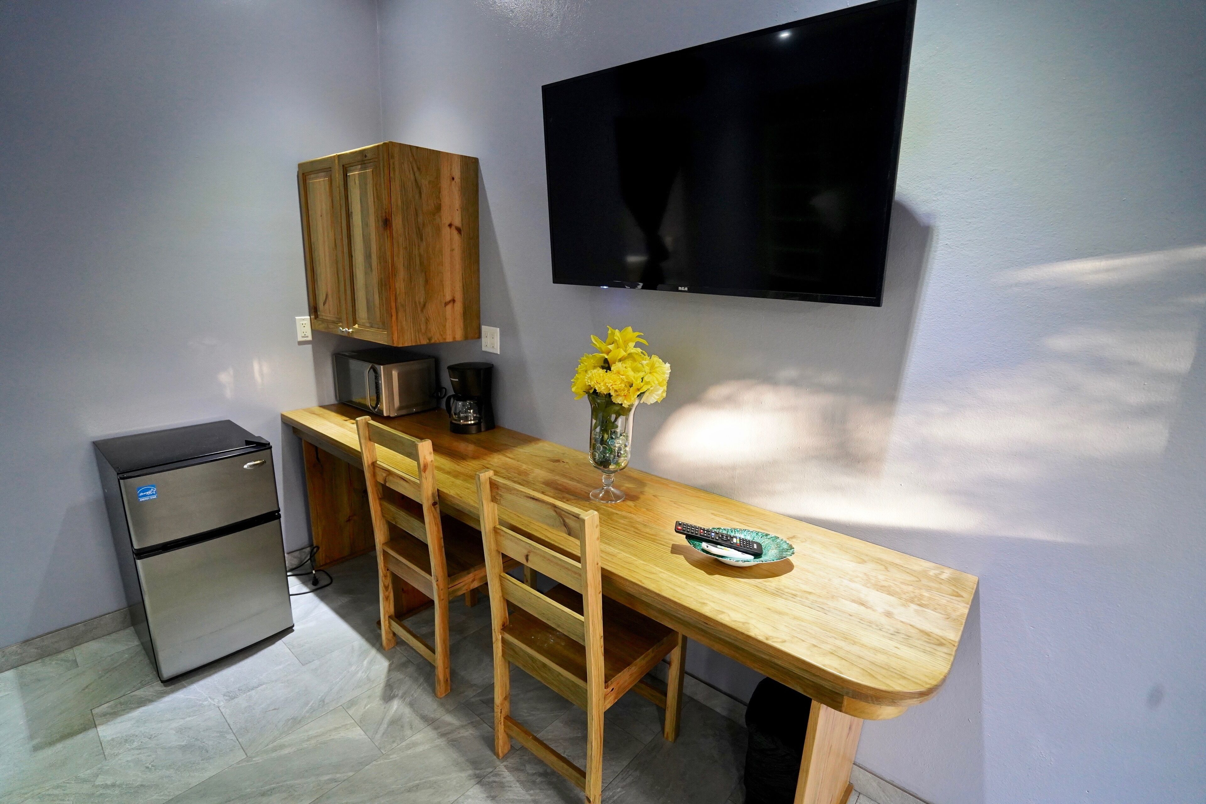 Cocina compacta con refrigerador, armarios de madera, televisor montado en la pared y mesa de comedor con sillas.