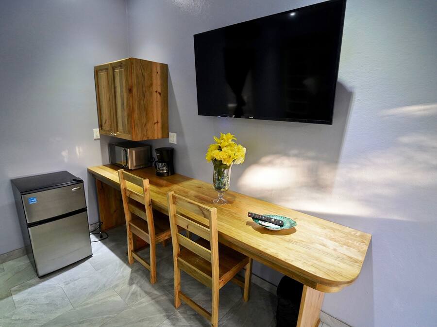 Cocina compacta con refrigerador, armarios de madera, televisor montado en la pared y mesa de comedor con sillas.