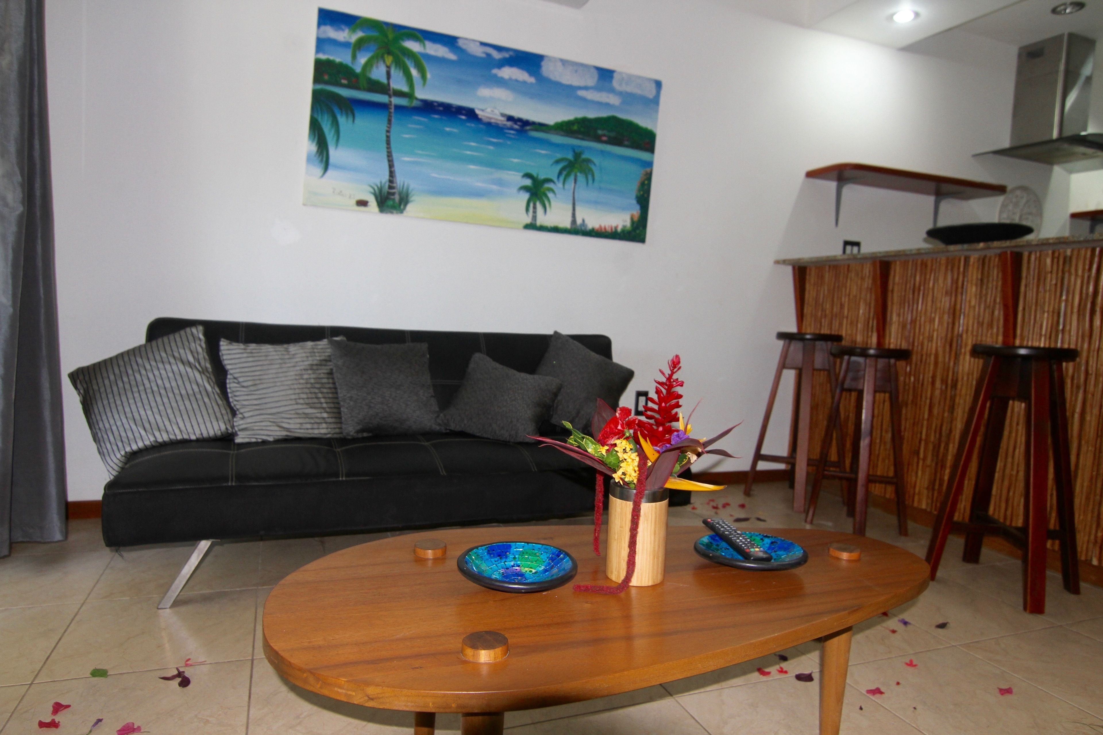 A living room with a black sofa, a wooden coffee table with decorative items, a painting of a beach scene, and a kitchen area with wooden stools.