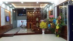 A hotel lobby with a wooden reception desk, a wall-mounted clock, and a large floral arrangement.