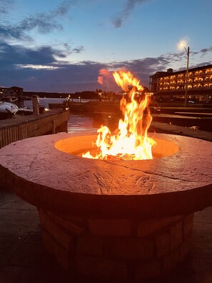 A circular stone fire pit with flames, surrounded by a waterfront area with boats and a building in the background. - Alexandria Bay, 1000 Islands Waterfront Cottage For Rent directly on the water, (Alexandria Bay)