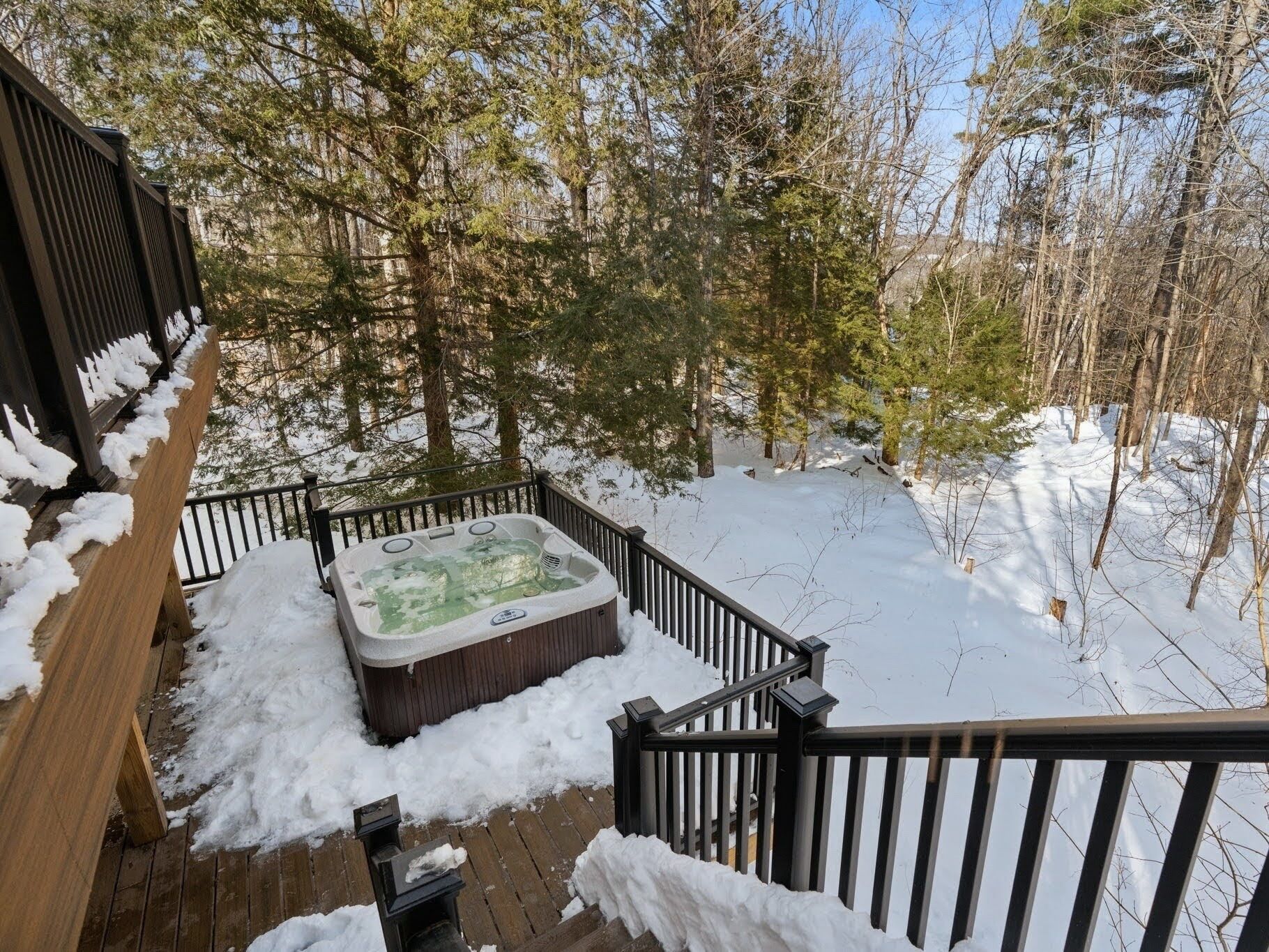 Un bain à remous sur une terrasse en bois, entouré de neige, avec une balustrade en bois et des arbres en arrière-plan.