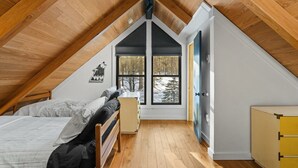Een slaapkamer met schuine wand, houten vloer, een bed, een dressoir en een raam met uitzicht op bomen.