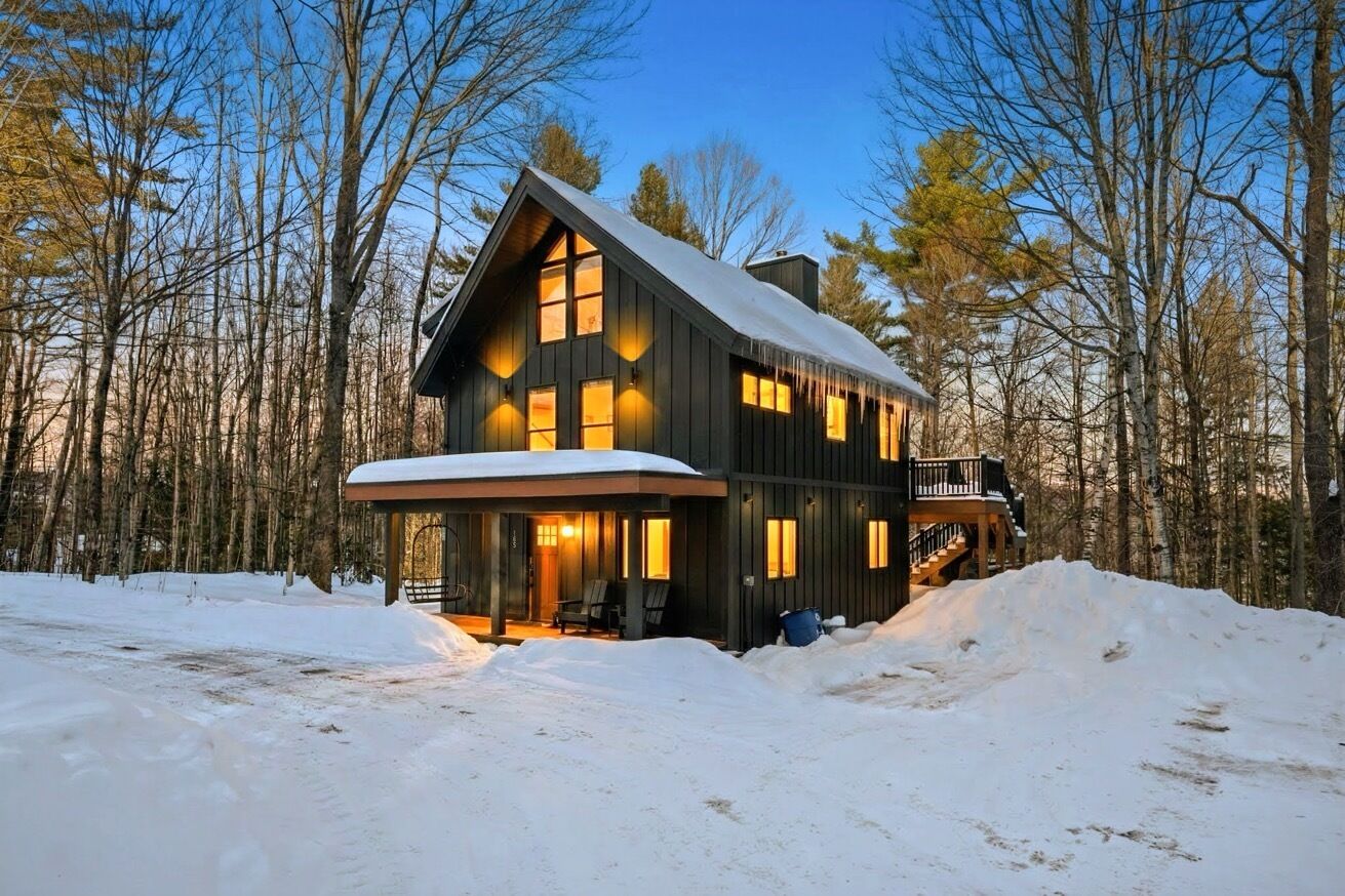 A large, dark wooden cabin with multiple windows and a covered porch, surrounded by a snowy landscape and trees.