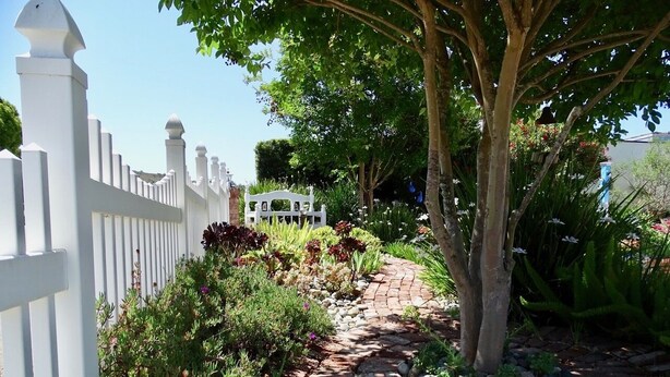 Un sendero en un jardín con cerca blanca, un árbol y diversas plantas.
