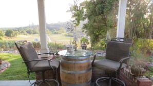 Un patio con dos sillas de mimbre y una mesa de barril de madera, rodeado de un jardín y vistas al campo.