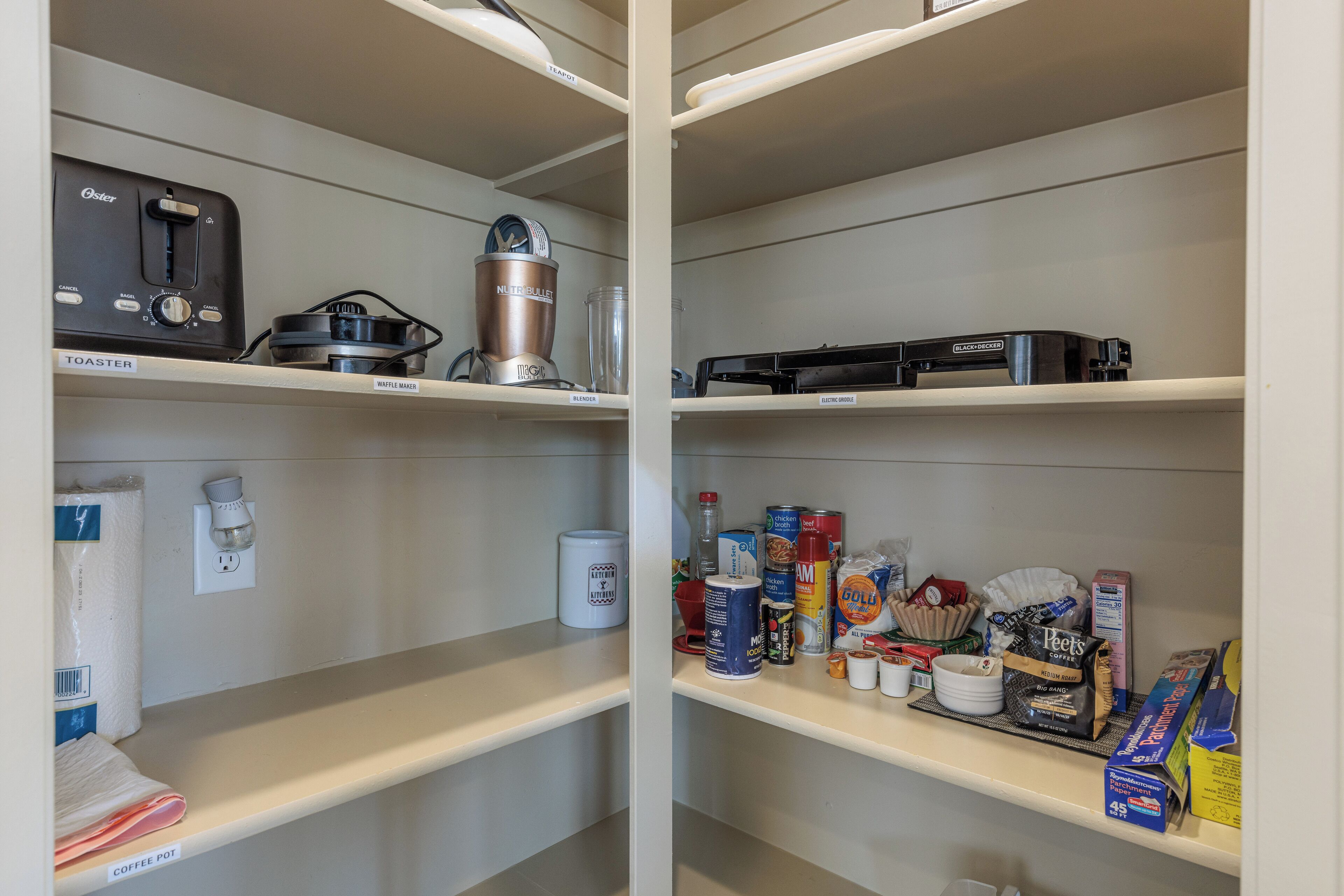 A kitchen with a toaster, waffle maker, coffee pot, and various food items on the shelves.